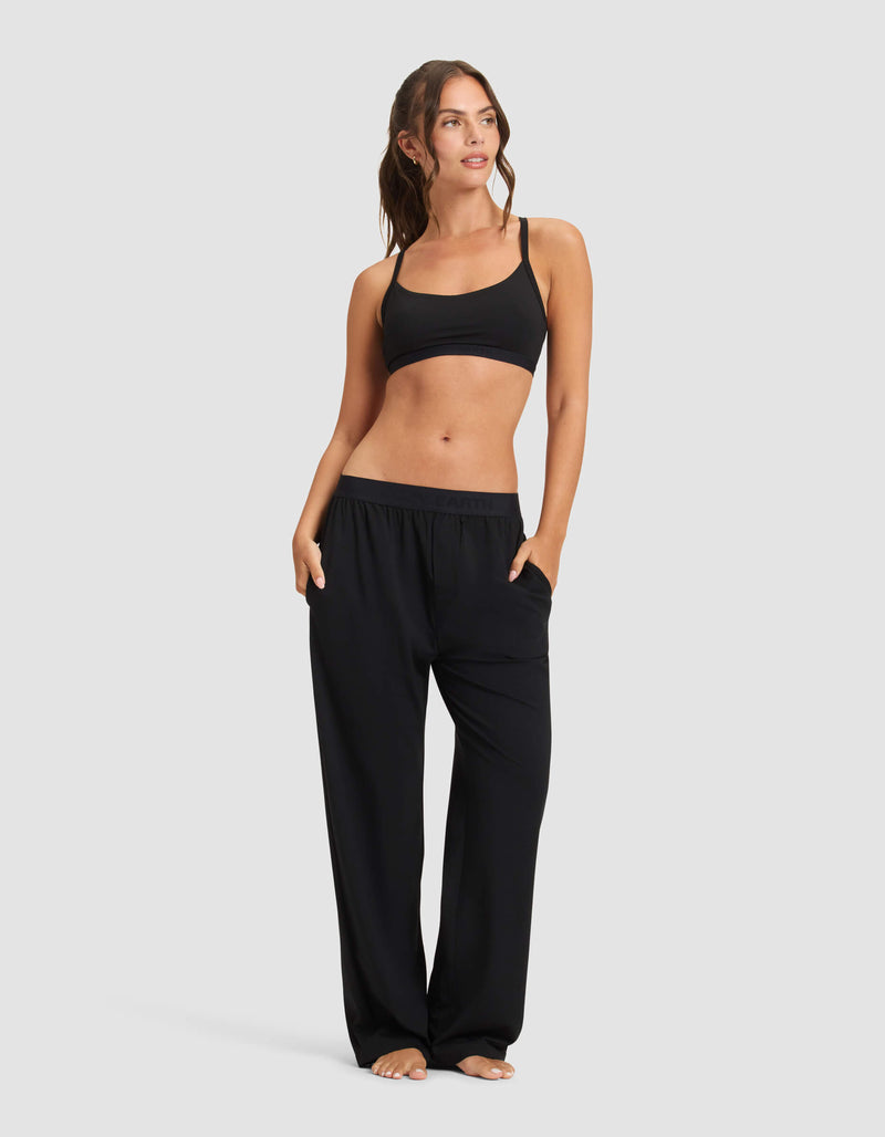 A woman with long brown hair in a loose ponytail stands barefoot against a plain background, wearing Cozy Earth’s Women's First Layer Pajama Pant in black and a black sports bra, hands in pockets, looking to the side.
