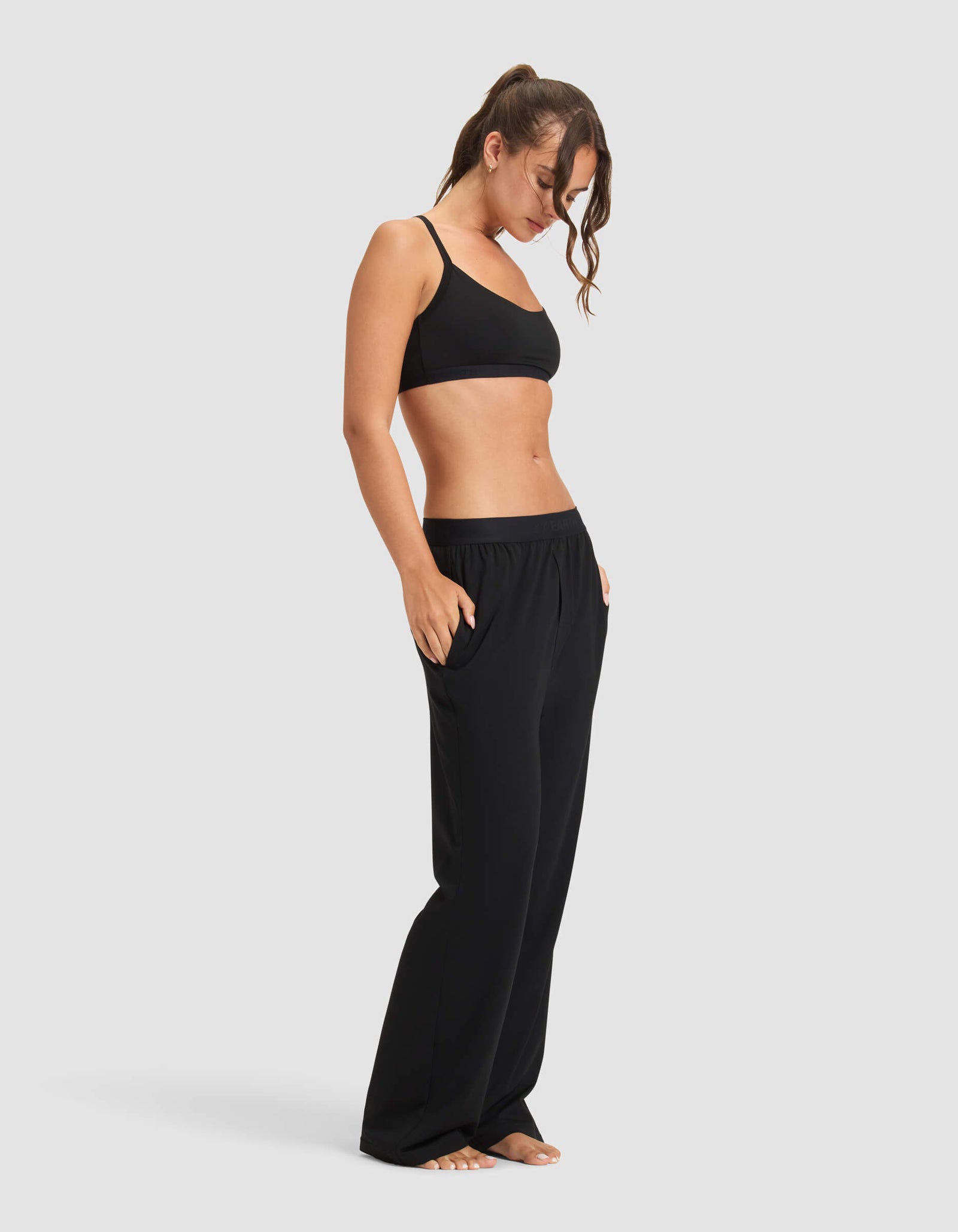 A woman stands barefoot against a plain background, wearing the Cozy Earth Women's First Layer Pajama Soft Bra & Pant Set in black, hands in pockets and looking down with a relaxed, casual posture.