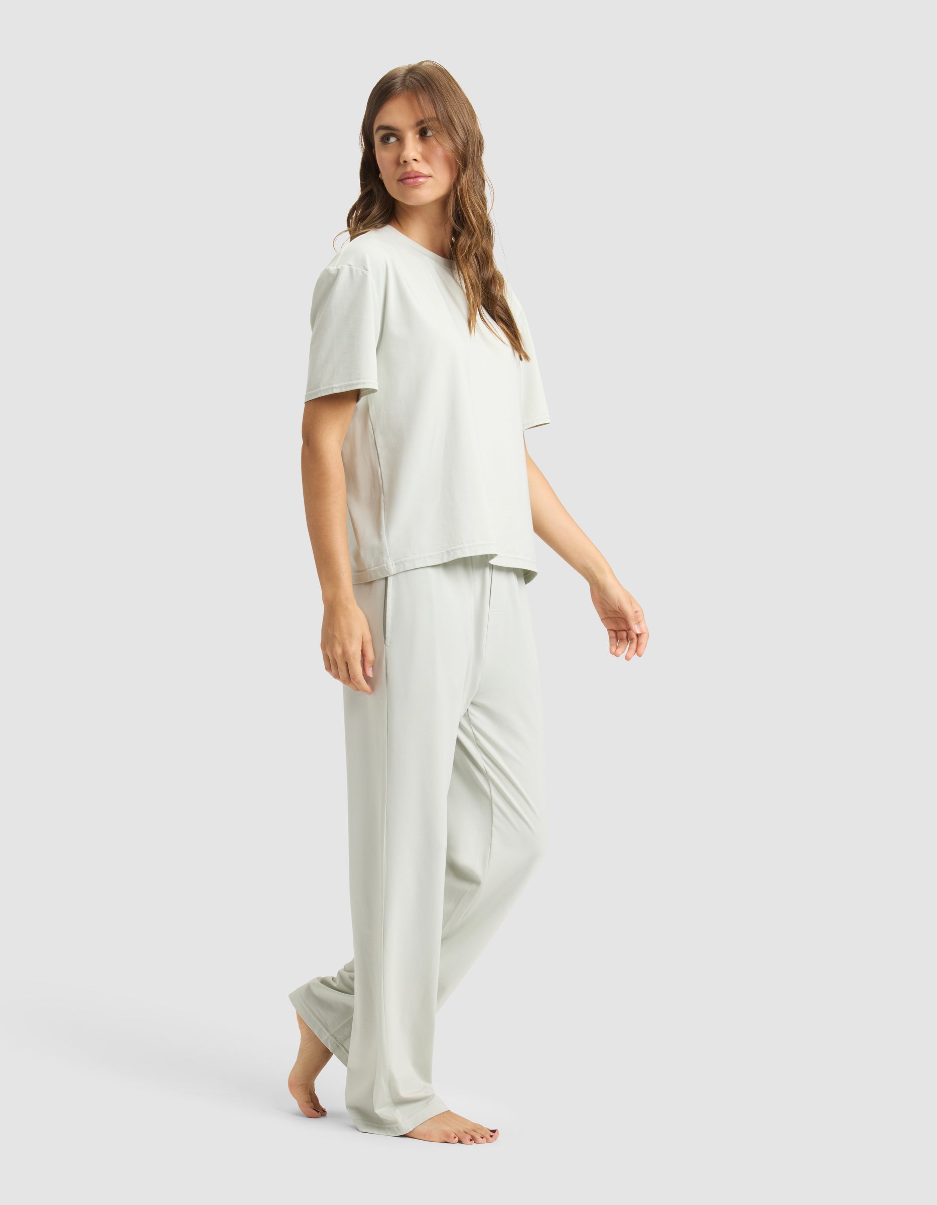 A woman with long brown hair wears the Cozy Earth Women's First Layer Pajama Short Sleeve Tee & Pant Set in light gray, featuring a loose fit and wide-leg pants, standing barefoot against a plain light background.|Color:Laurel