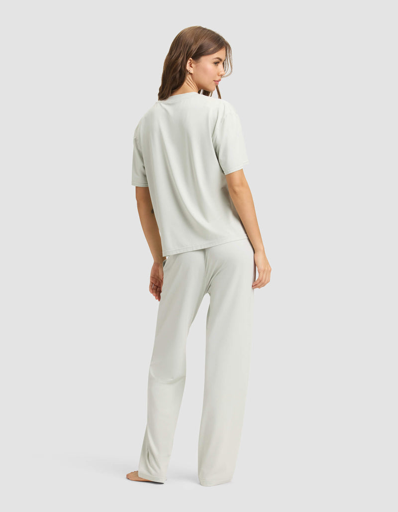 A woman stands barefoot and facing away, wearing the Cozy Earth Women's First Layer Pajama Short Sleeve Tee & Pant Set in light grey, against a plain light background.