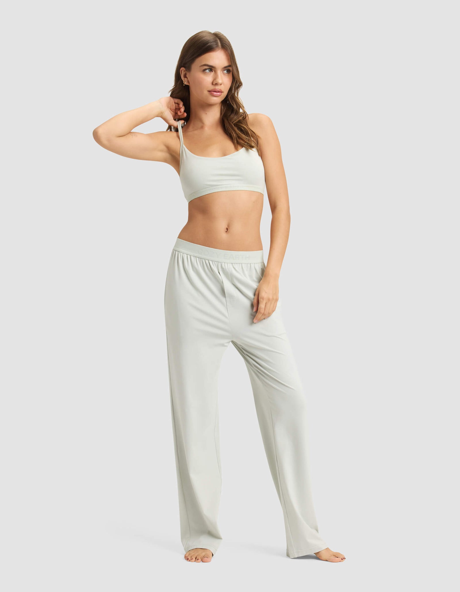 A woman with long wavy hair stands barefoot against a plain background, wearing the Cozy Earth Women's First Layer Pajama Soft Bra & Pant Set in light gray, with one hand resting on her head.