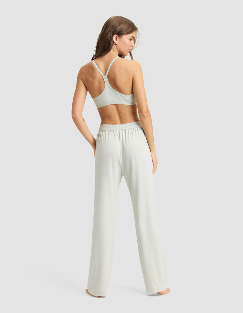 A woman stands barefoot, facing away, wearing the Cozy Earth Women's First Layer Pajama Soft Bra & Pant Set in a light color against a plain background.