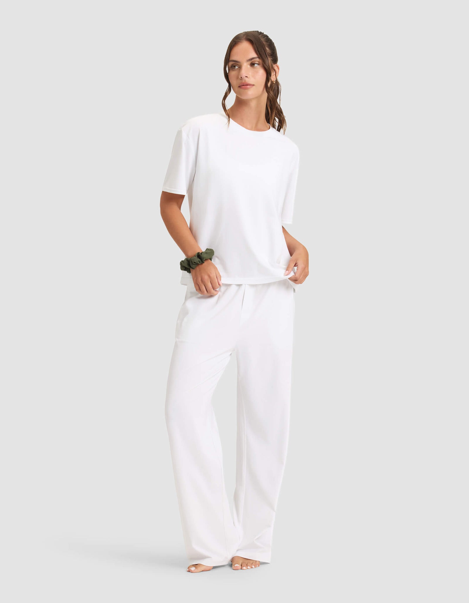A woman models the Cozy Earth Women's First Layer Pajama Short Sleeve Tee & Pant Set in white, barefoot against a plain background, with a dark scrunchie on her wrist and her hair in a loose ponytail.
