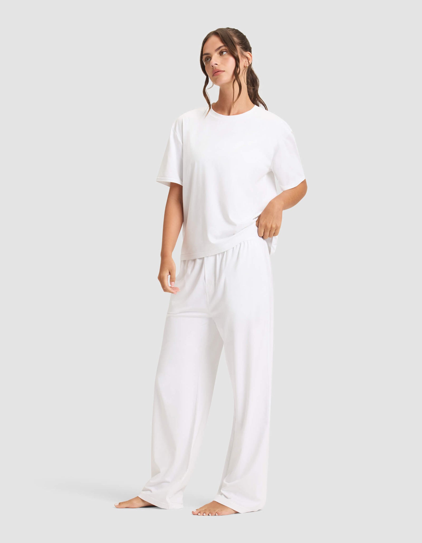 A woman with long, wavy brown hair stands barefoot against a plain background, wearing the Cozy Earth Women's First Layer Pajama Short Sleeve Tee & Pant Set in white. She gazes slightly to the side.