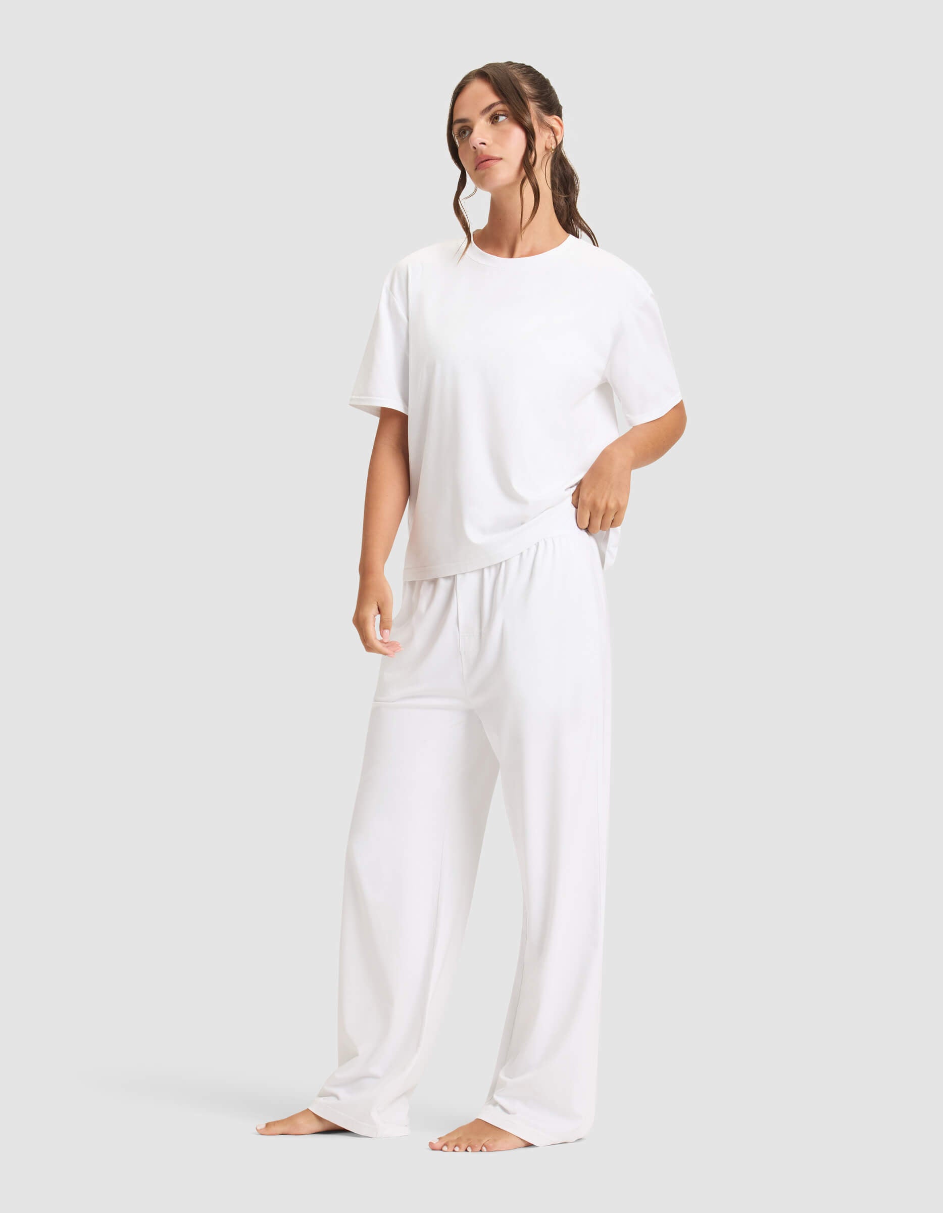 A woman with long, wavy brown hair stands barefoot against a plain background, wearing the Cozy Earth Women's First Layer Pajama Short Sleeve Tee & Pant Set in white. She gazes slightly to the side.|Color:White