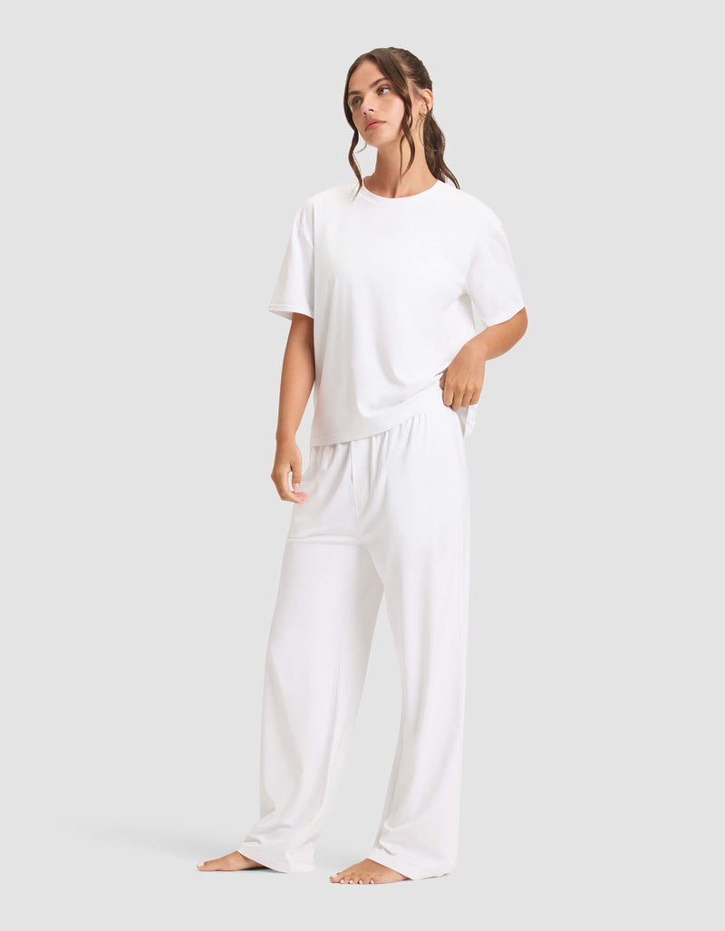 A woman with long, wavy brown hair stands barefoot against a plain background, wearing the Cozy Earth Women's First Layer Pajama Short Sleeve Tee & Pant Set in white. She gazes slightly to the side.