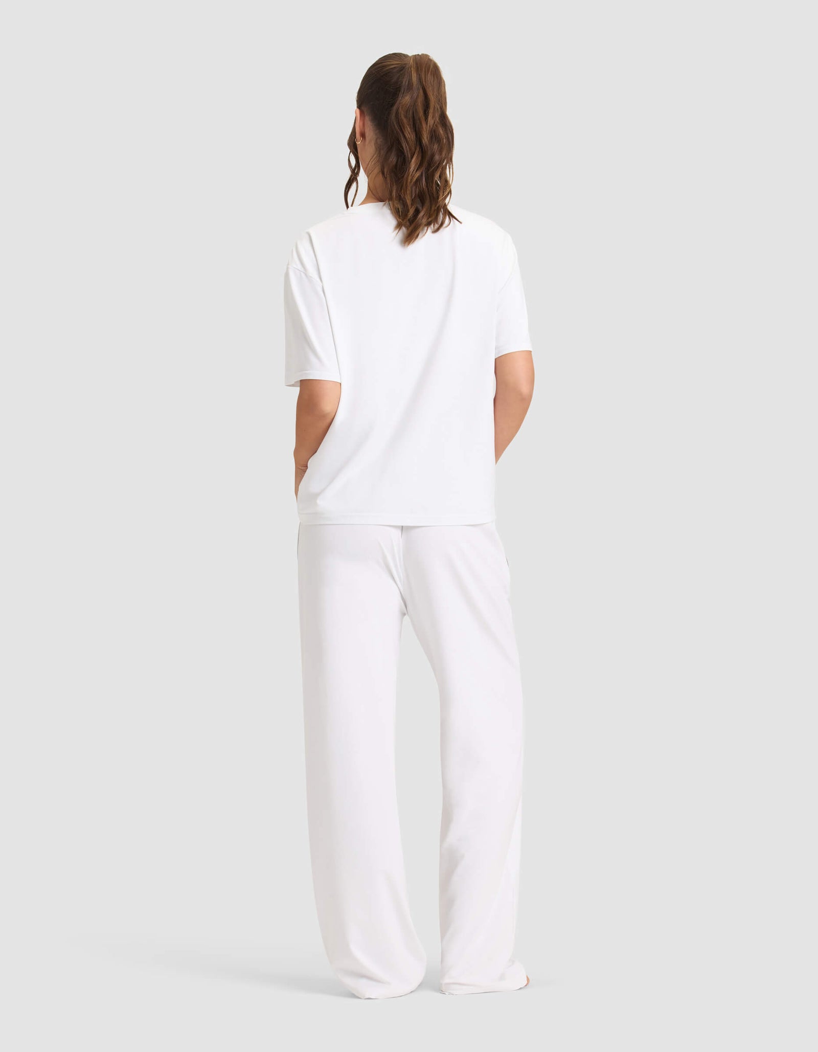A woman with wavy brown hair in a ponytail faces away from the camera, wearing the Cozy Earth Women's First Layer Pajama Short Sleeve Tee & Pant Set in white against a plain light gray background.