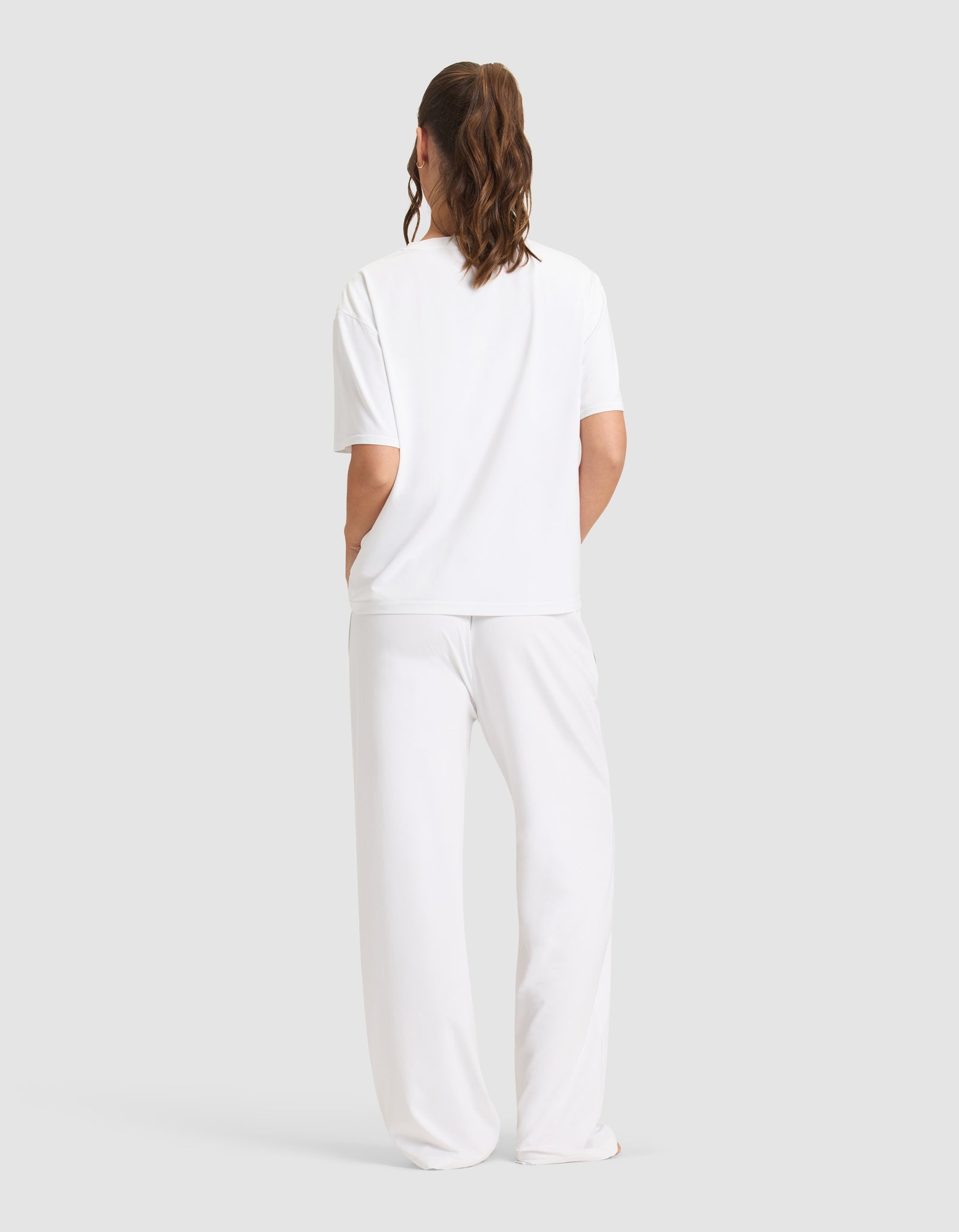 A person stands facing away, wearing the Cozy Earth Women's First Layer Pajama Short Sleeve Tee & Pant Set in white against a plain light gray background.