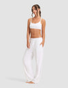 A woman models the Cozy Earth Women's First Layer Pajama Soft Bra & Pant Set, barefoot against a plain light gray background, with loose brown hair, hands in her pockets, and looking slightly to the side.|Color:White
