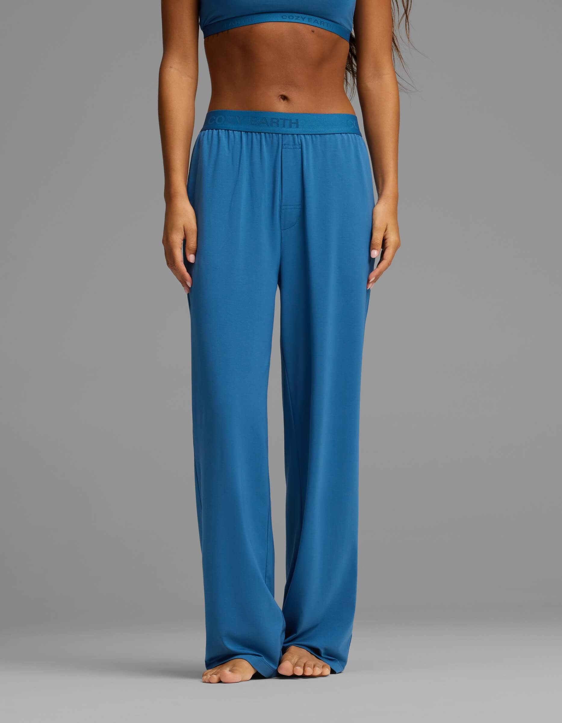 A person stands barefoot, wearing Cozy Earth’s Women’s First Layer Pajama Pant in blue with a matching top. The image is cropped from the shoulders down, with hands resting at the sides against a plain gray background.