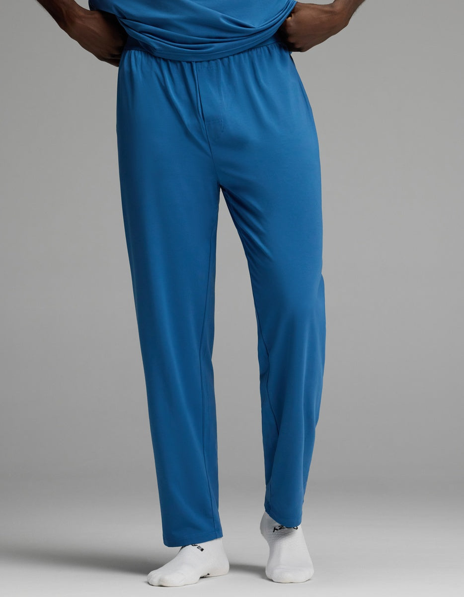 A person is shown from the shoulders down wearing Cozy Earth Men’s First Layer Pajama Pant in blue with white socks, standing against a plain gray background.