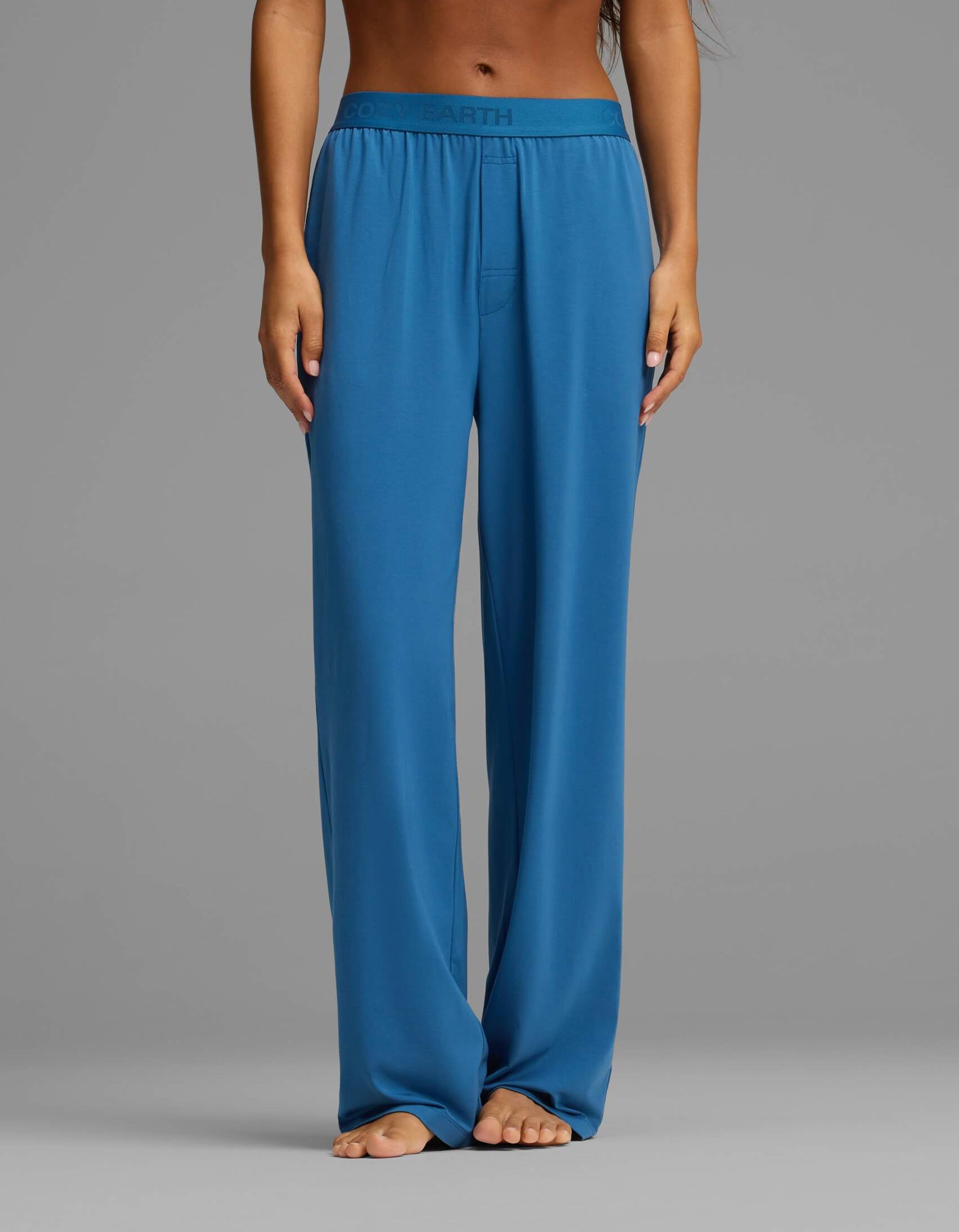 A person is shown barefoot, wearing the Cozy Earth Women’s First Layer Pajama Short Sleeve Tee & Pant Set in blue. The image focuses on the lower torso and legs, highlighting the relaxed, high-waisted pants with an elastic waistband against a plain gray background.