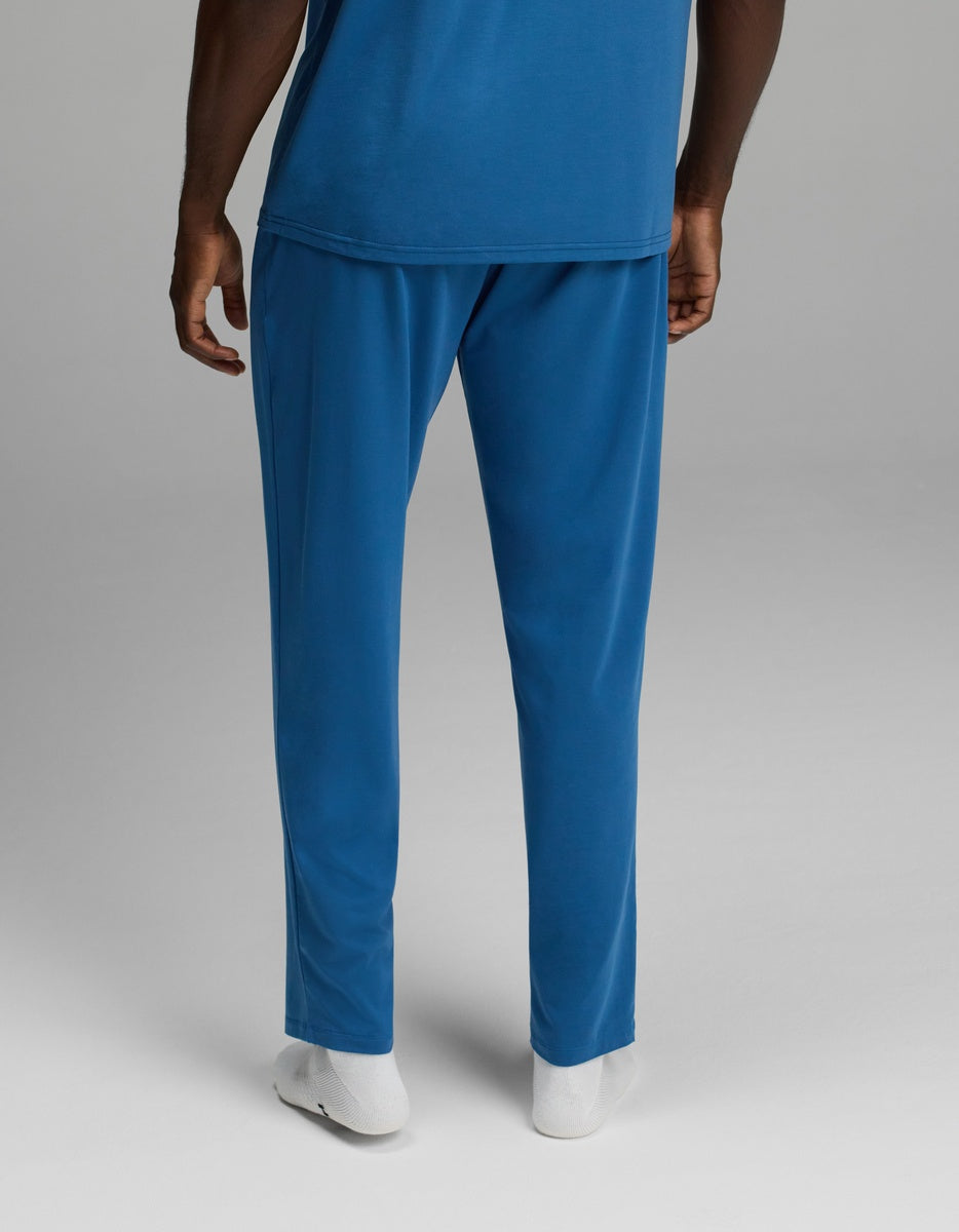 A person is shown from the lower torso down, wearing Cozy Earth’s Men’s First Layer Pajama Pant and a blue shirt. They are also wearing white socks, standing against a plain gray background with only their pants, part of their arms, and legs visible.|Color:Anchor