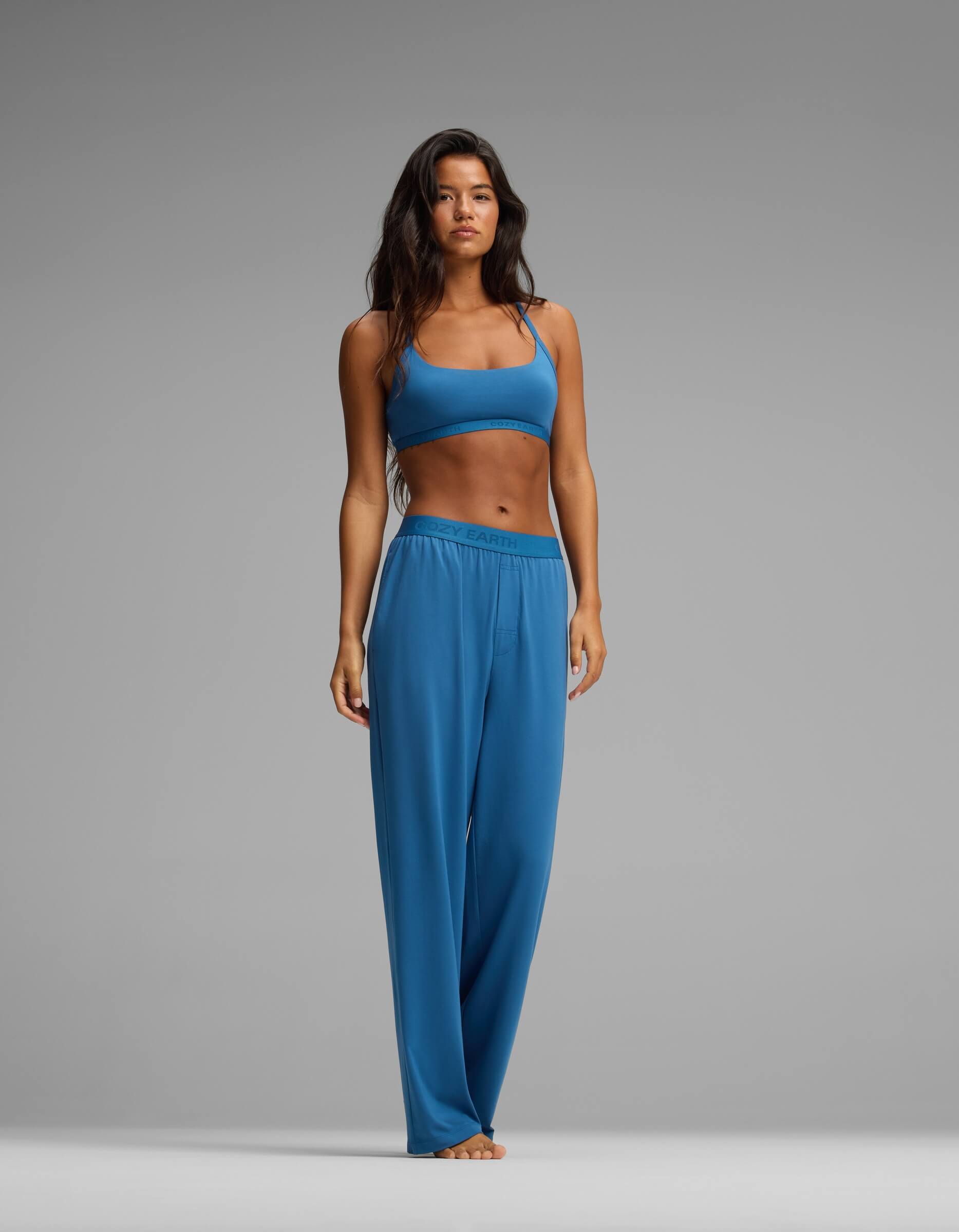 A woman stands barefoot against a gray background, wearing the Cozy Earth Women’s First Layer Pajama Pant in blue with a matching blue sports bra. She has long dark hair and a neutral expression, her arms relaxed at her sides.