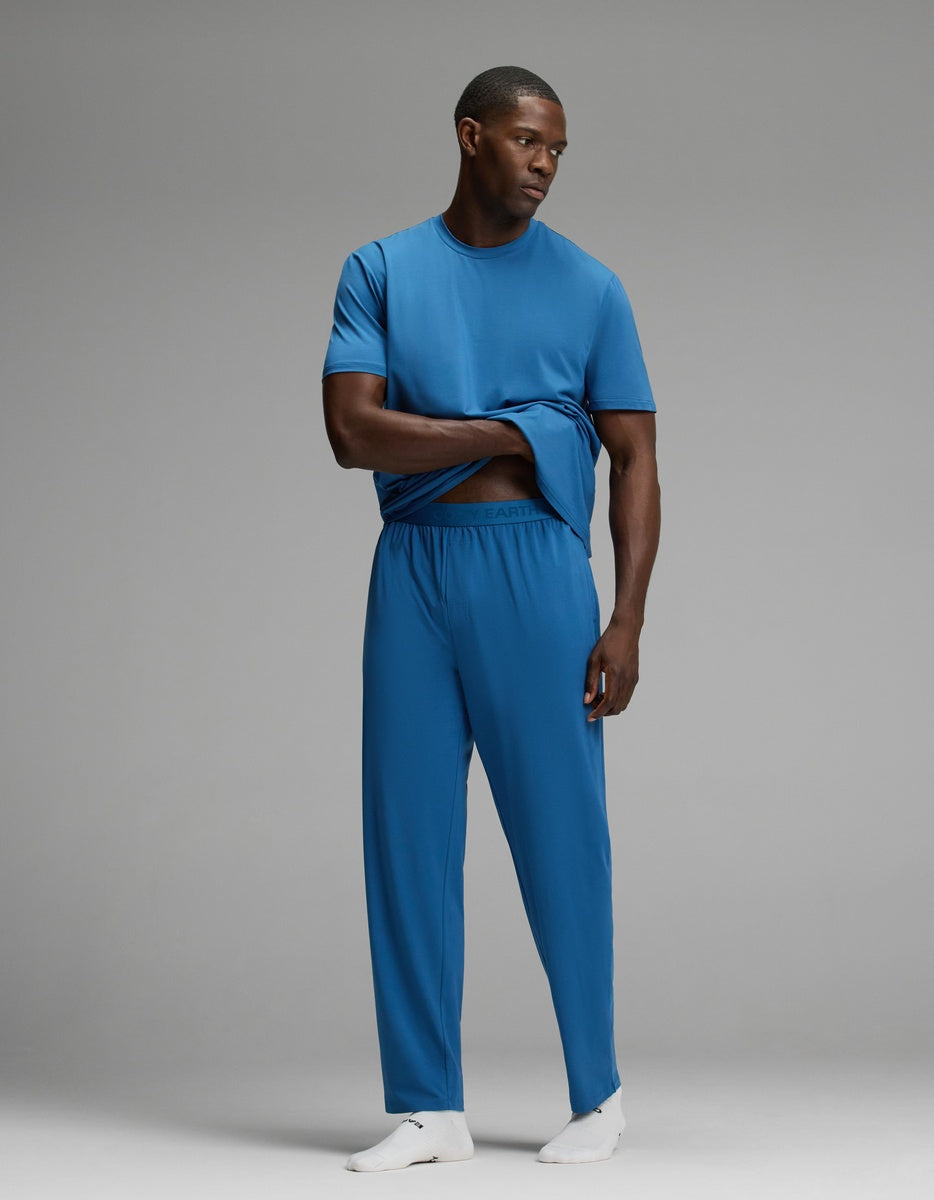 A man stands on a plain backdrop, wearing Cozy Earth’s Men’s First Layer Pajama Pant in blue with a matching blue t-shirt. He lifts his shirt slightly with one hand and also wears white socks.