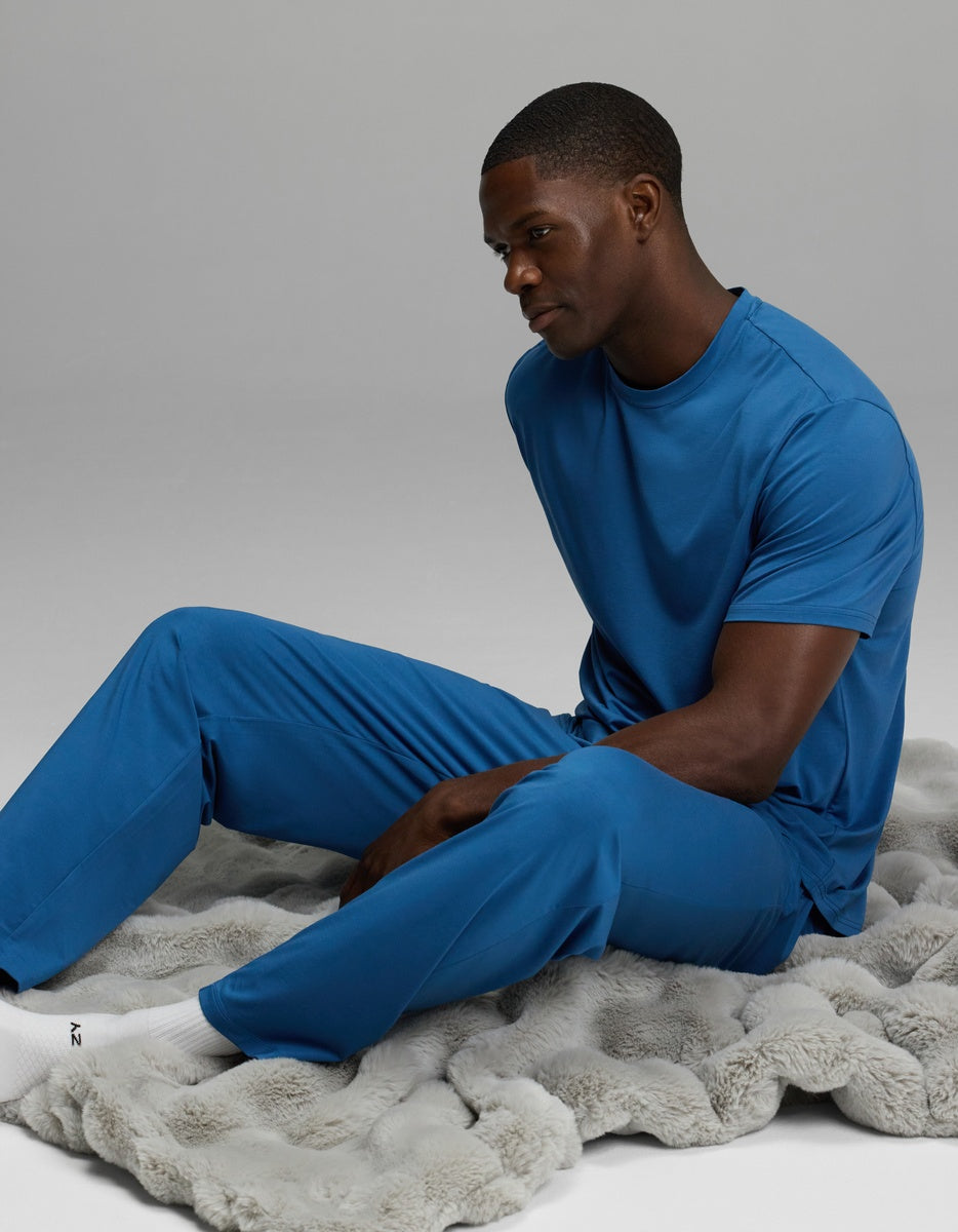 A man dressed in Cozy Earth’s Men’s First Layer Pajama Pant sits on a soft, gray textured blanket against a plain background, looking thoughtfully to the side.