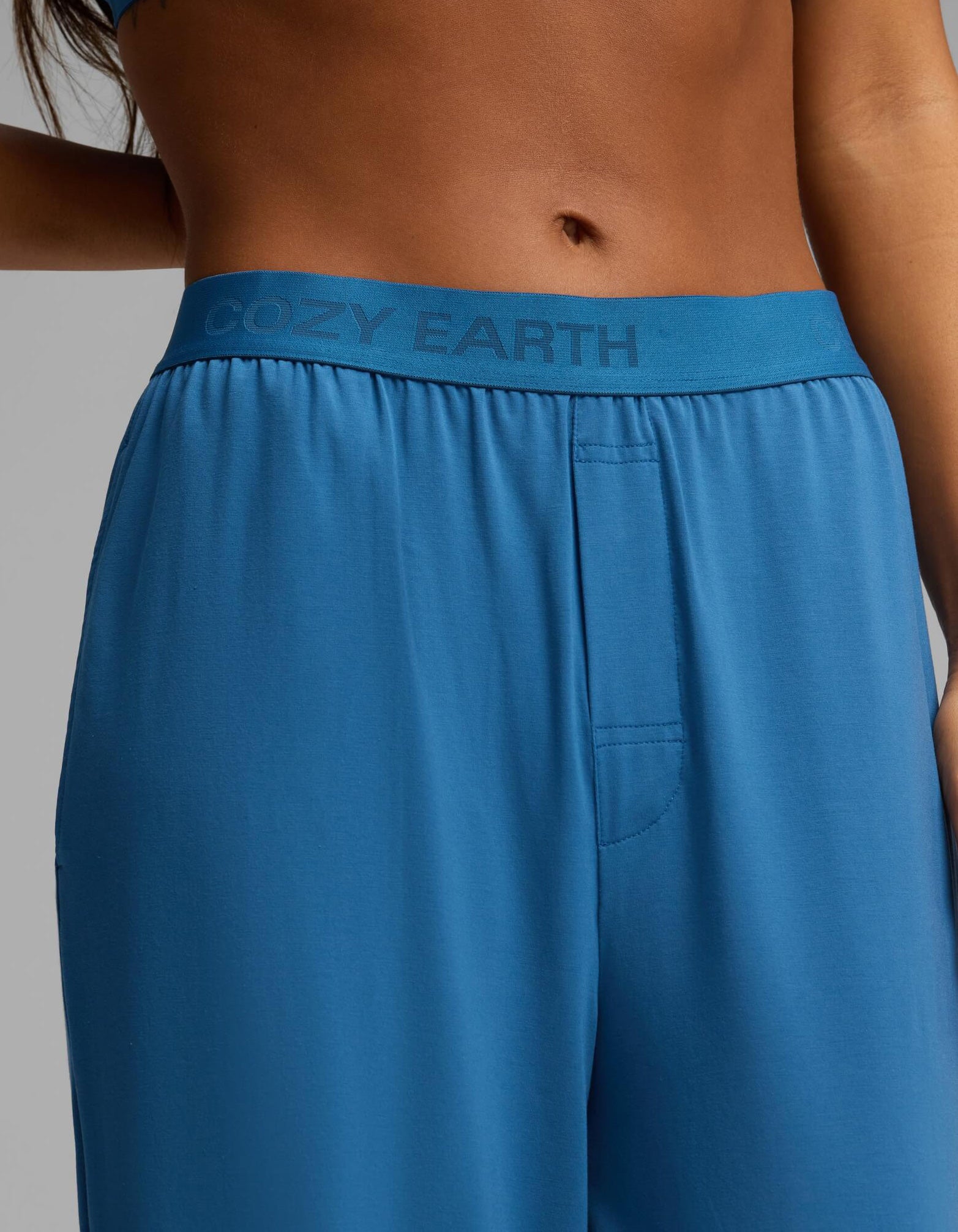 A close-up shows the lower torso and part of one arm of a person wearing the Cozy Earth Women’s First Layer Pajama Short Sleeve Tee & Pant Set in blue, with “COZY EARTH” visible on the waistband.