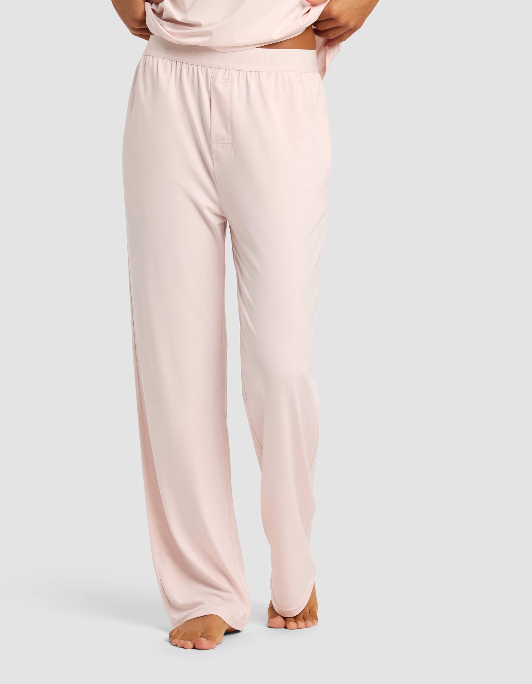 A woman models Cozy Earth's Women's First Layer Pajama Pant in light pink, standing barefoot on a light gray background with hands on hips and the pants gently grazing the floor.