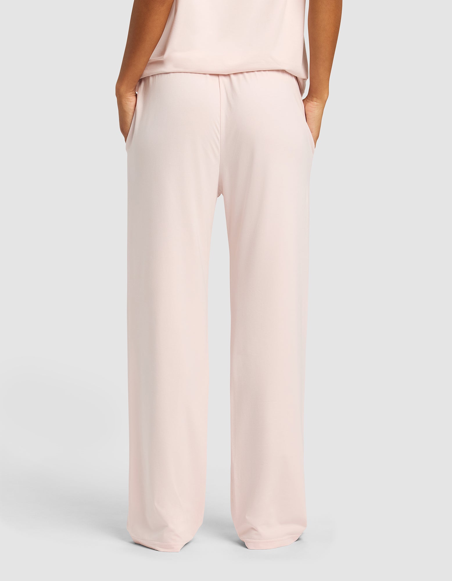 A woman wearing Cozy Earth Women's First Layer Pajama Pants in light pink, hands in pockets, is shown from the back against a plain light gray background.|Color:Ballet