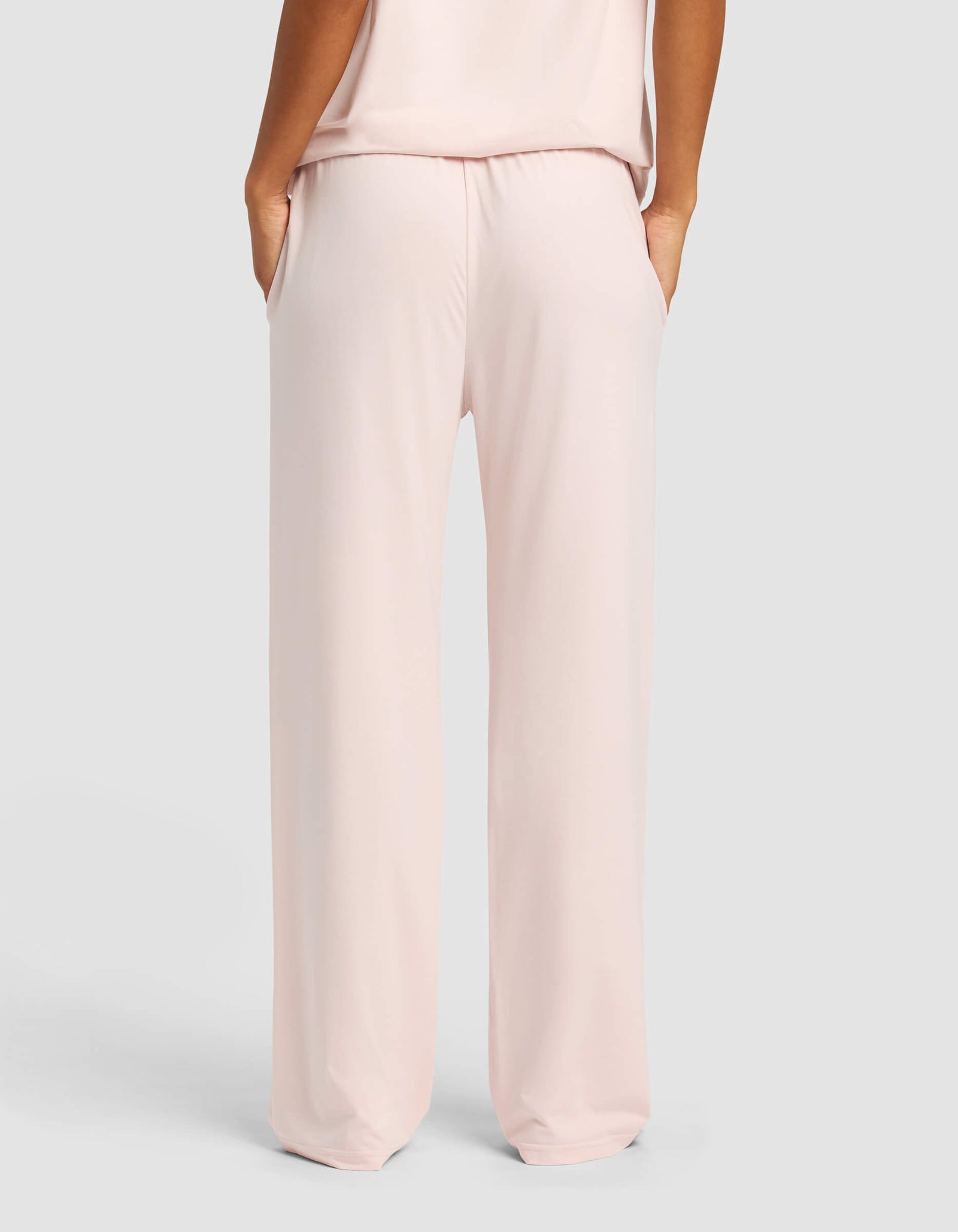 A woman wearing Cozy Earth Women's First Layer Pajama Pants in light pink, hands in pockets, is shown from the back against a plain light gray background.