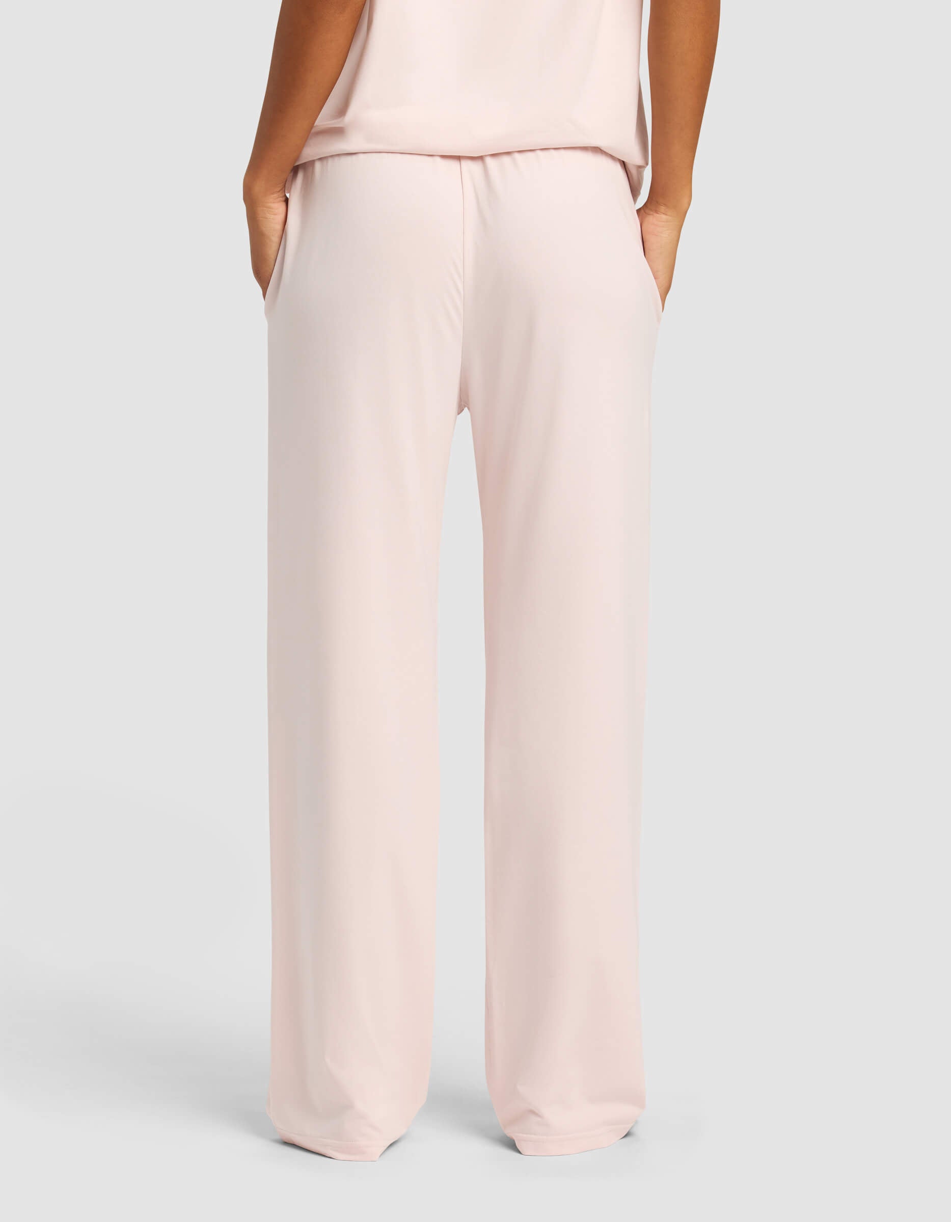 A woman wearing Cozy Earth Women's First Layer Pajama Pants in light pink, hands in pockets, is shown from the back against a plain light gray background.|Color:Ballet