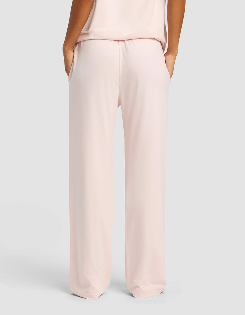 A woman wearing Cozy Earth Women's First Layer Pajama Pants in light pink, hands in pockets, is shown from the back against a plain light gray background.