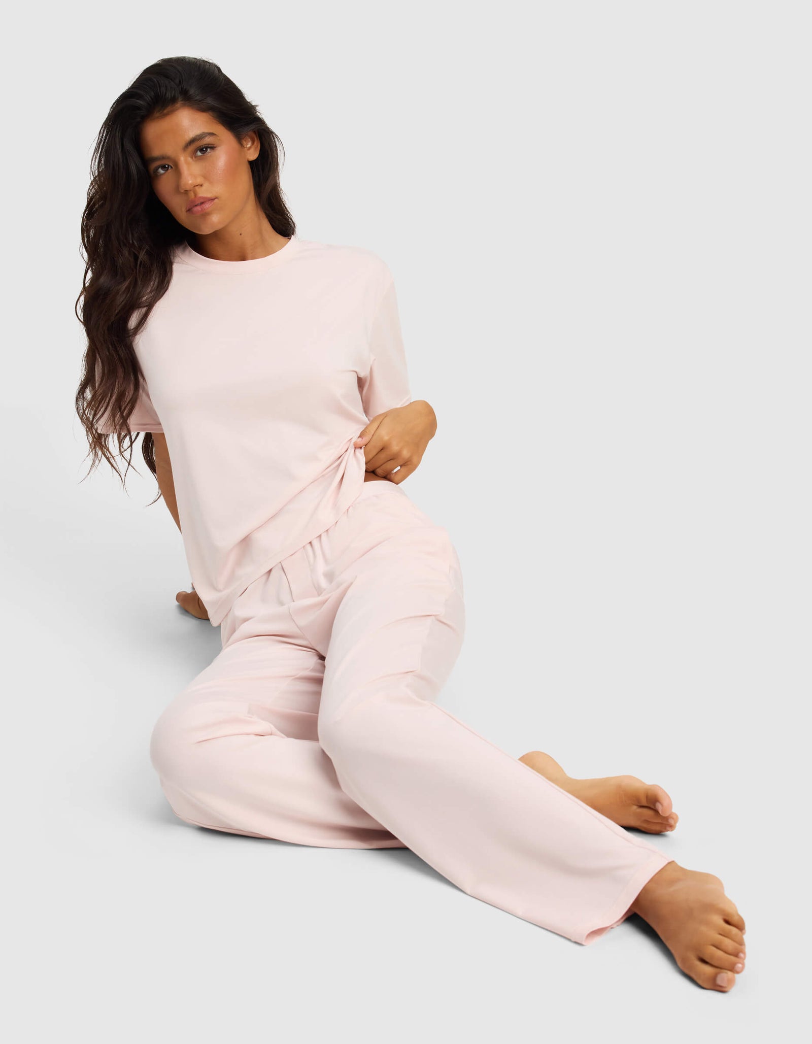 A woman with long dark hair sits on the floor against a light background, wearing the Cozy Earth Women's First Layer Pajama Short Sleeve Tee & Pant Set in light pink, looking at the camera with a relaxed expression.