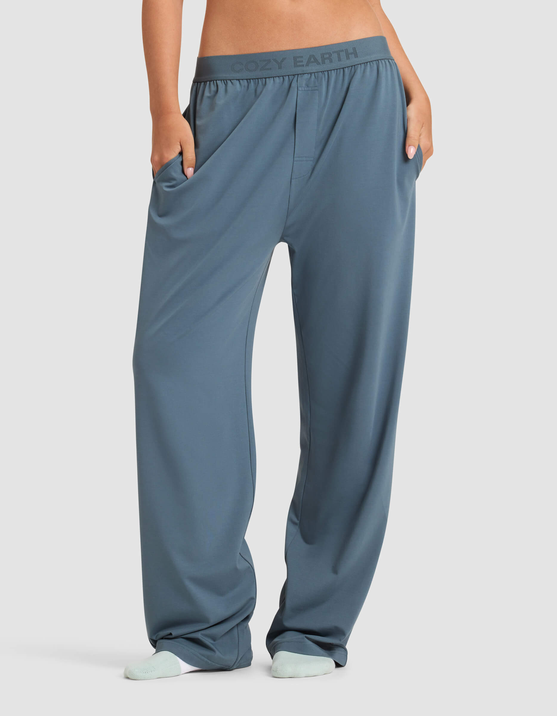 A person models the Cozy Earth Women's First Layer Pajama Pant—loose-fitting blue pants with pockets and a branded waistband—shown from the waist down, hands in pockets, paired with light-colored socks.