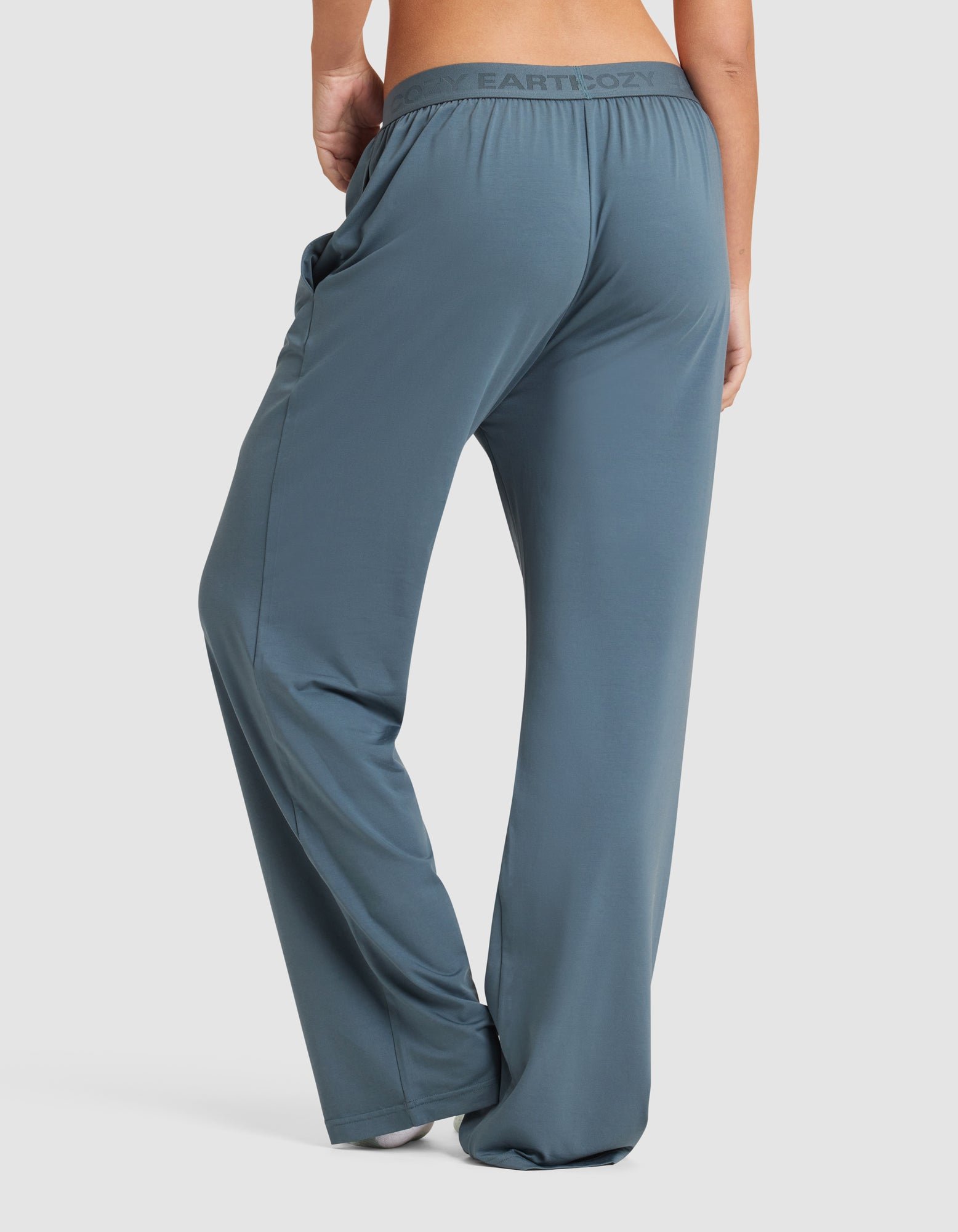 A woman stands barefoot against a plain white background, shown from the back wearing Cozy Earth Women's First Layer Pajama Pant in a loose-fitting, high-waisted blue-gray style.