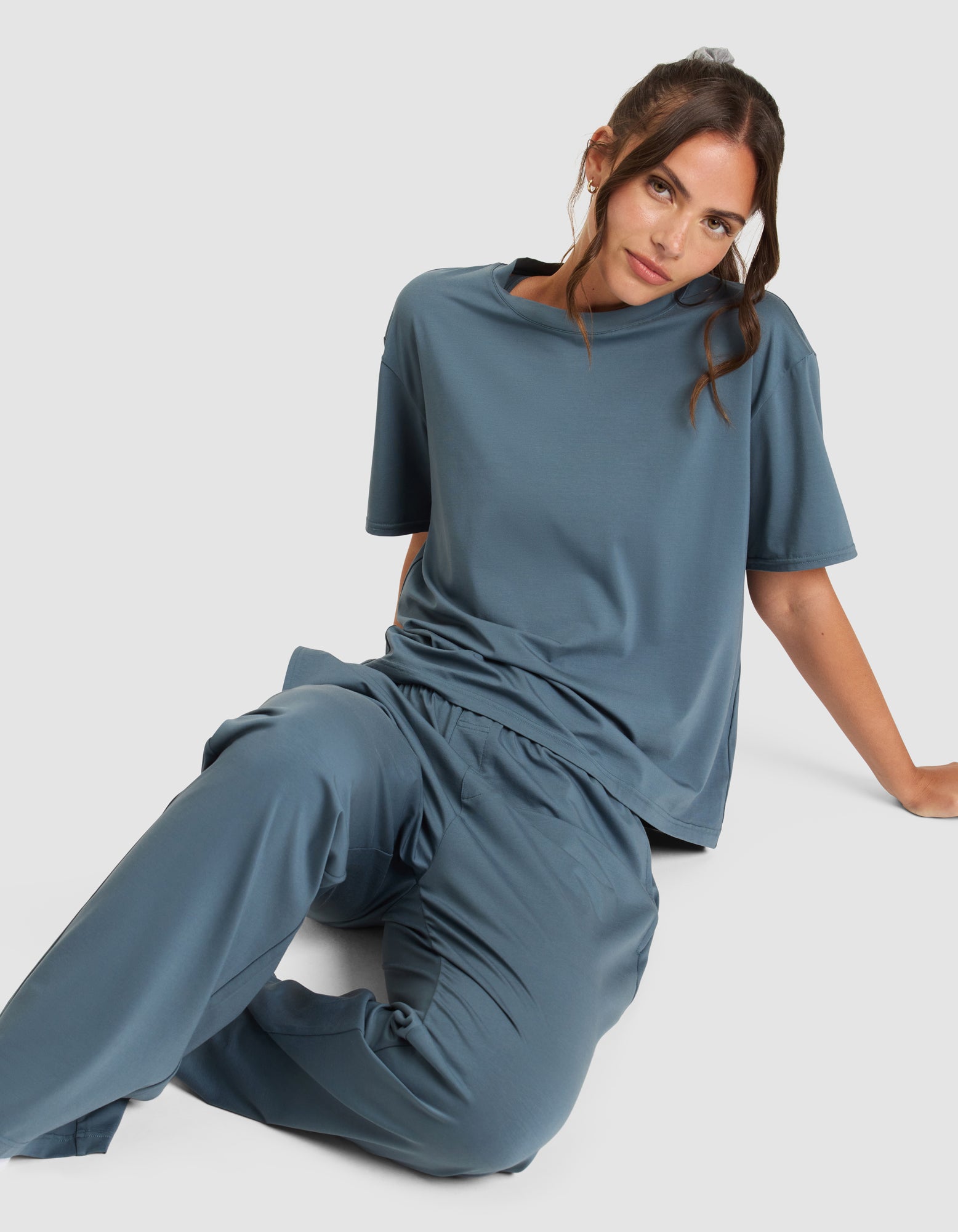 A woman with brown hair sits on the floor against a plain light background, wearing Cozy Earth's Women's First Layer Pajama Pant in blue with a matching loose short-sleeve top, looking at the camera with a relaxed expression.