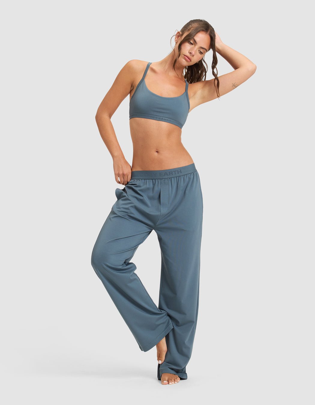 A woman models the Cozy Earth Women's First Layer Pajama Soft Bra & Pant Set in blue, barefoot against a light gray background, with one hand on her head and the other in her pocket.