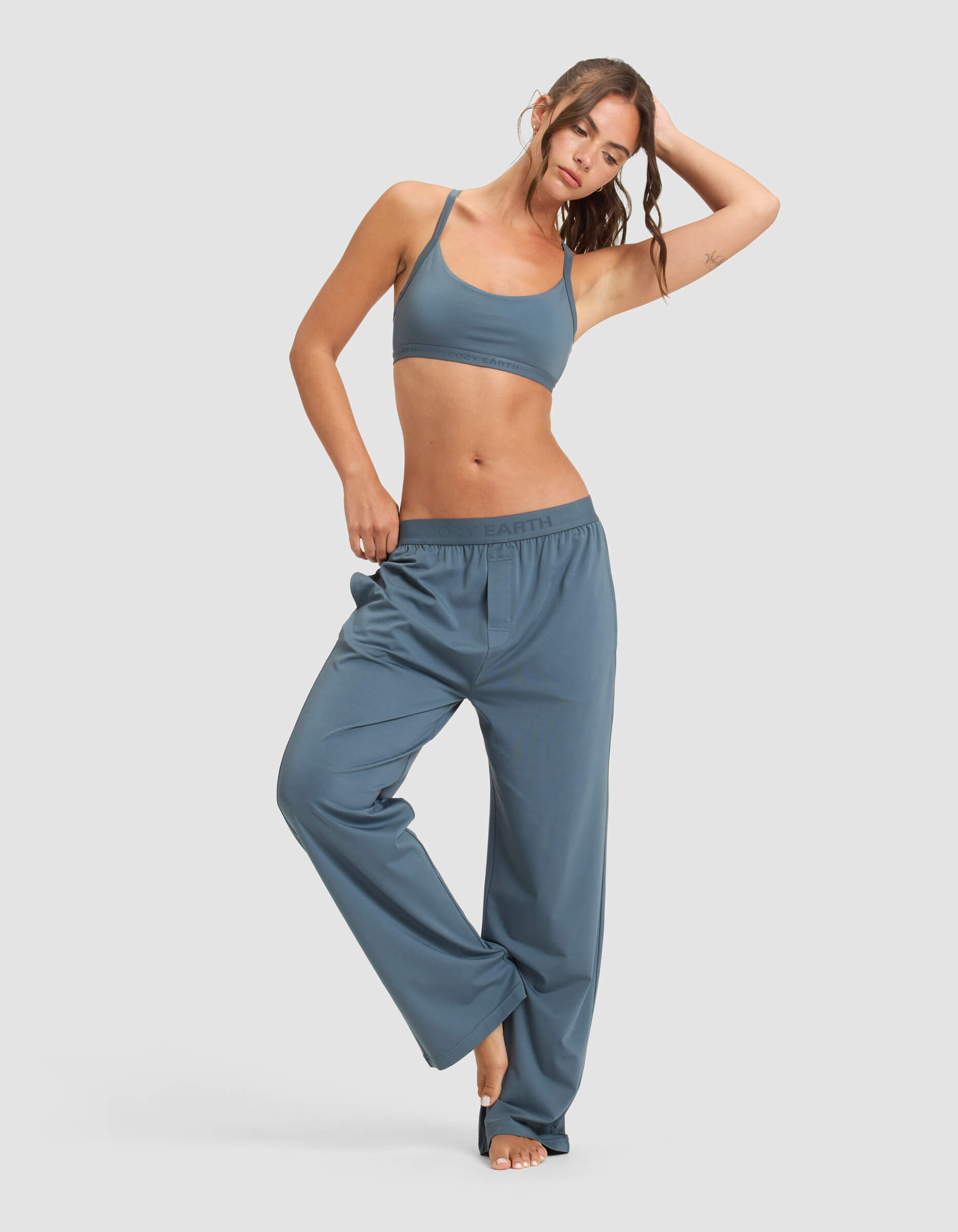 A woman models the Cozy Earth Women's First Layer Pajama Soft Bra & Pant Set—blue, loose, high-waisted pants and a matching bra—barefoot, balancing on one foot with her hand on her head against a plain light background.