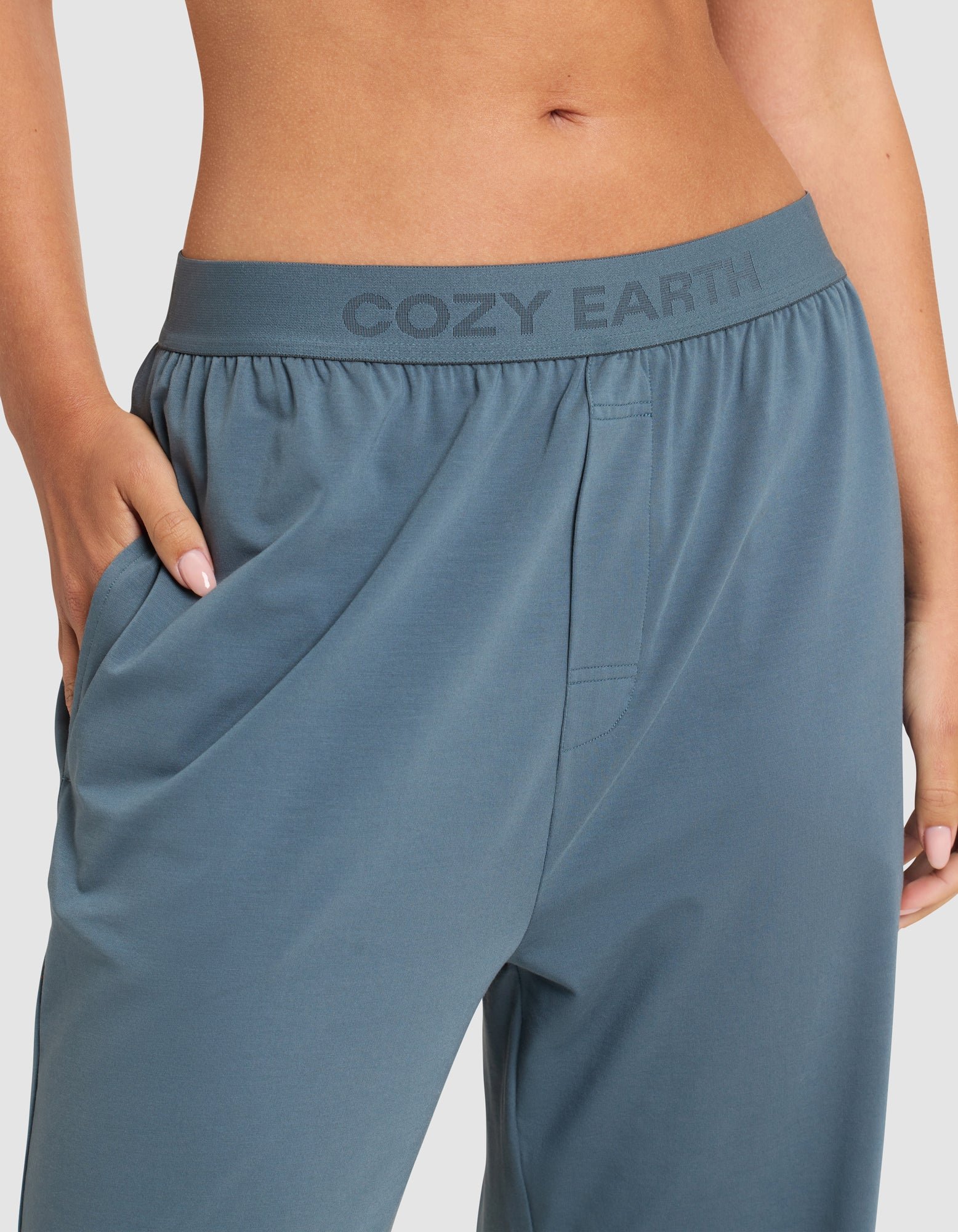 Close-up of a person wearing Cozy Earth Women's First Layer Pajama Pants with the brand name on the waistband. Their hand rests in the right pocket, and their midriff is visible against a plain background.
