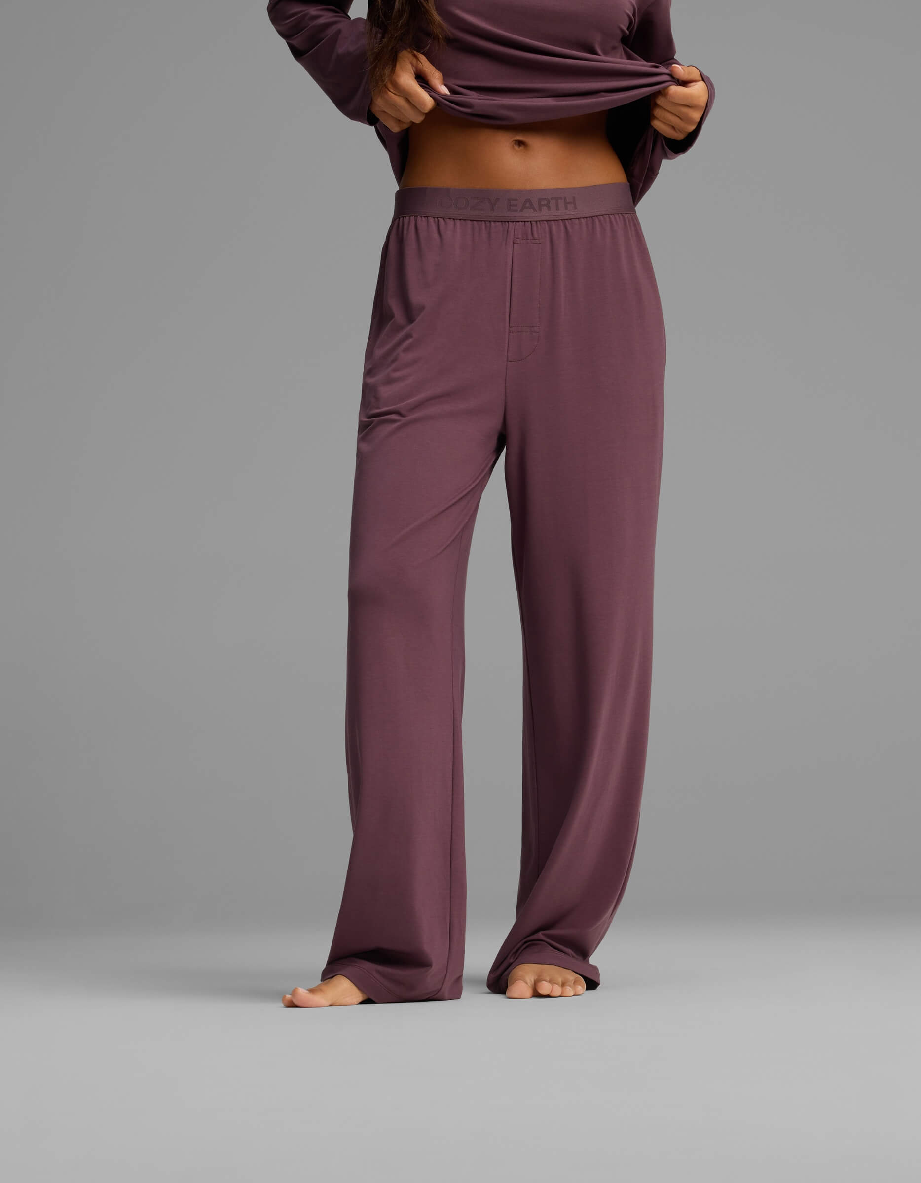 A person wears Cozy Earth’s Women’s First Layer Pajama Pant in mauve, standing barefoot against a plain gray background. The top of the person’s head is not visible in the image.