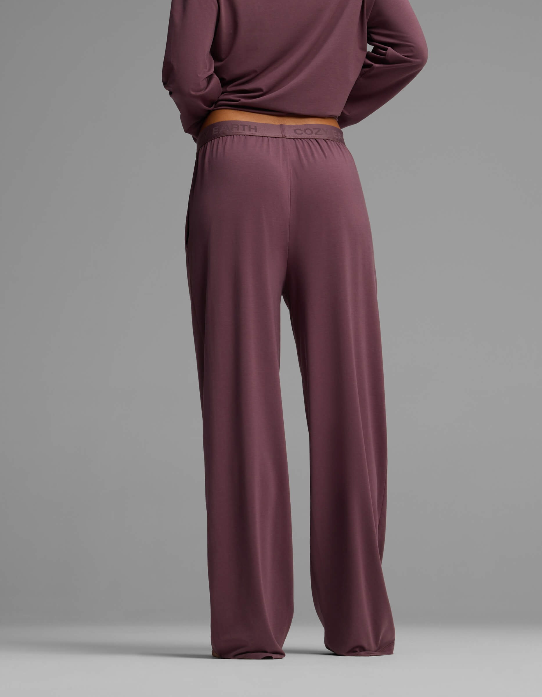 A woman is seen from the back wearing Cozy Earth’s Women’s First Layer Pajama Pant in a loose mauve fit, paired with a matching long-sleeve top, set against a plain gray background.