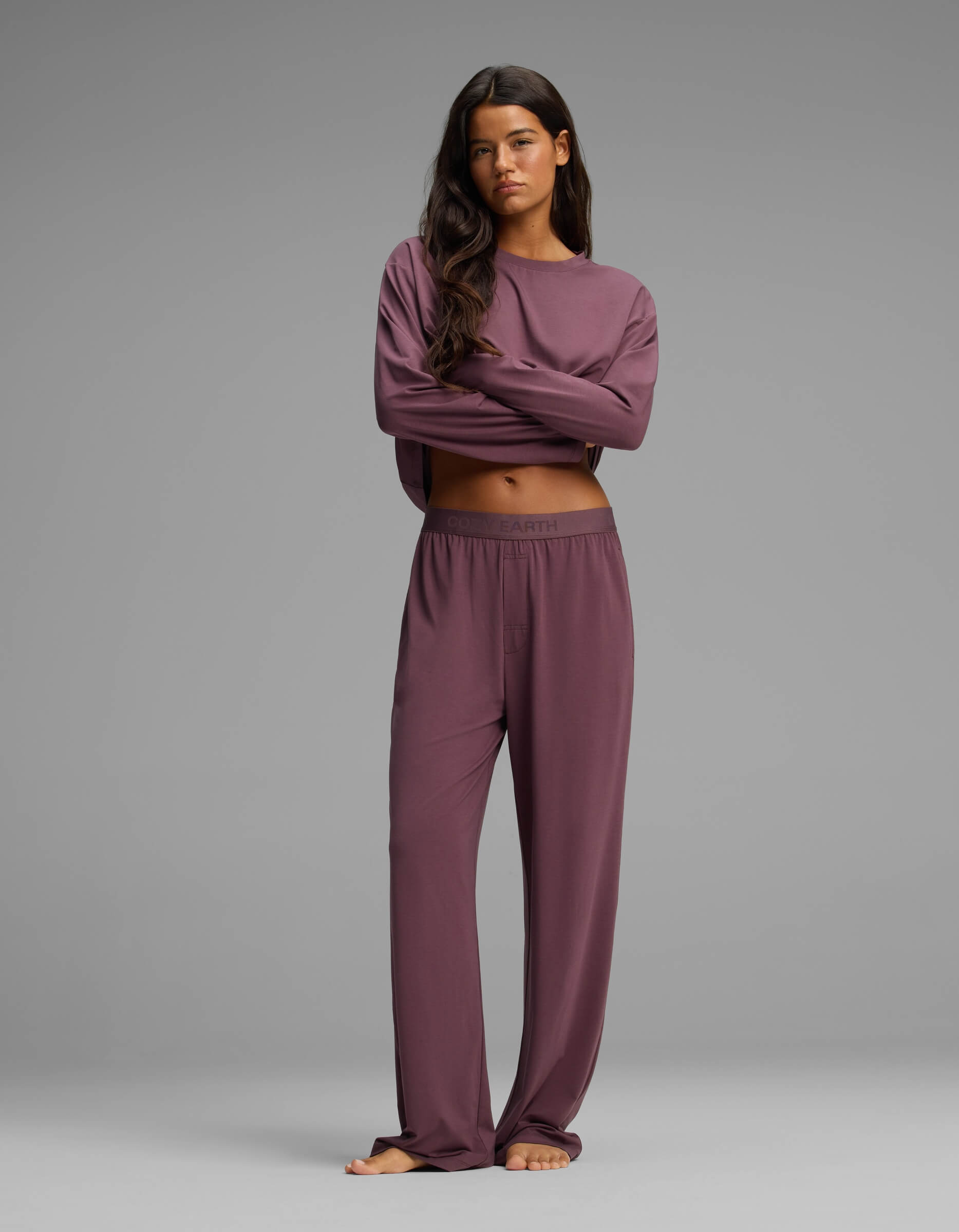 A woman stands barefoot with arms crossed, wearing Cozy Earth’s Women’s First Layer Pajama Pant in a loose-fitting, long-sleeved mauve set against a plain gray background.