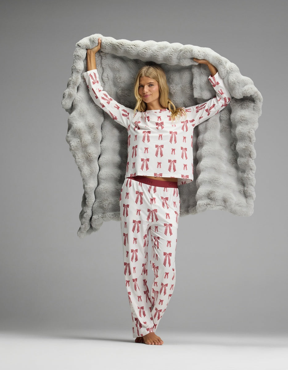 A woman wearing white Women’s First Layer Pajama Pant and a long-sleeved top with a red bow pattern, holding a large grey faux fur blanket over her shoulders.