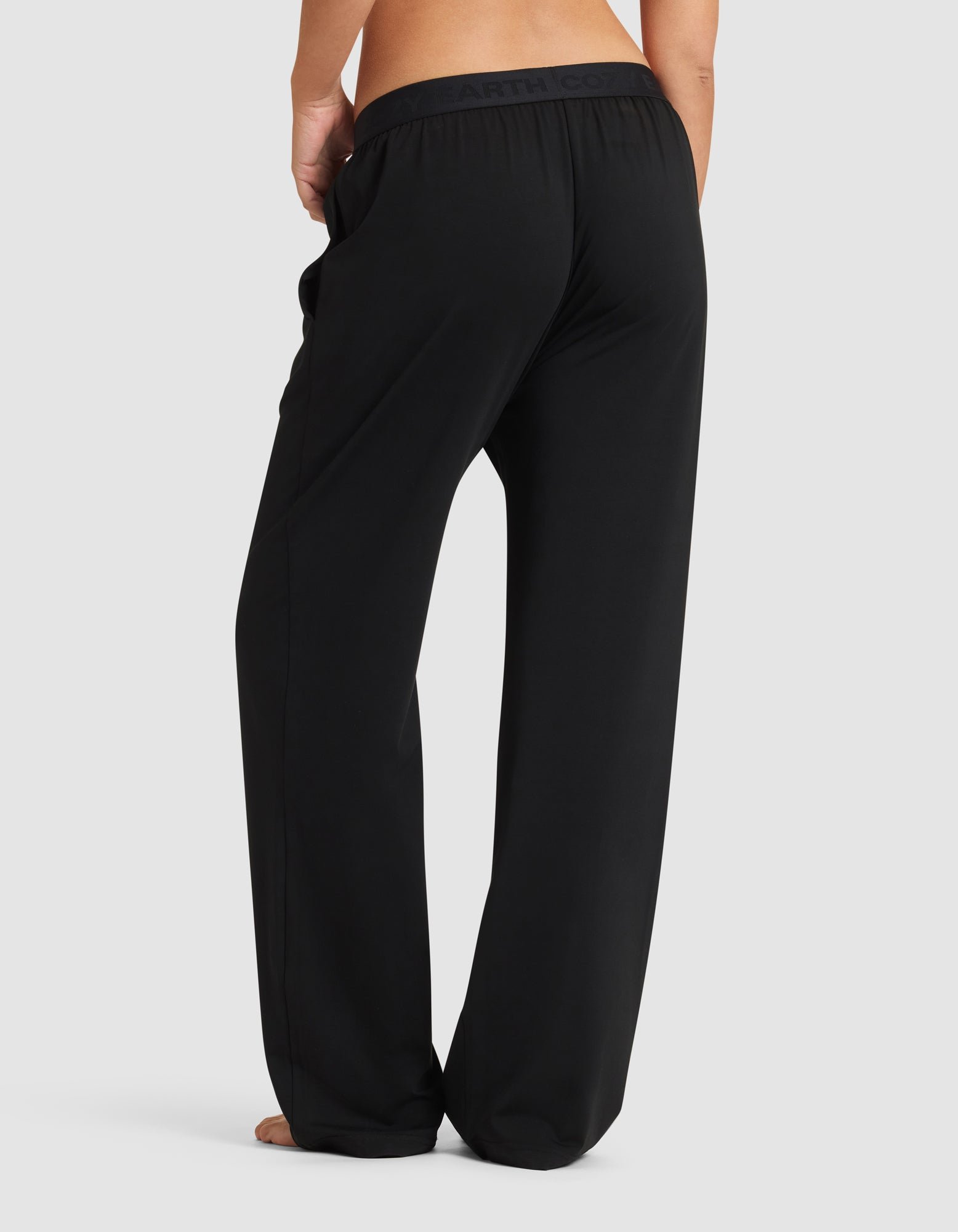 A woman models Cozy Earth’s Women's First Layer Pajama Pant in black, standing barefoot and facing away from the camera against a simple light background.|Color:Jet Black