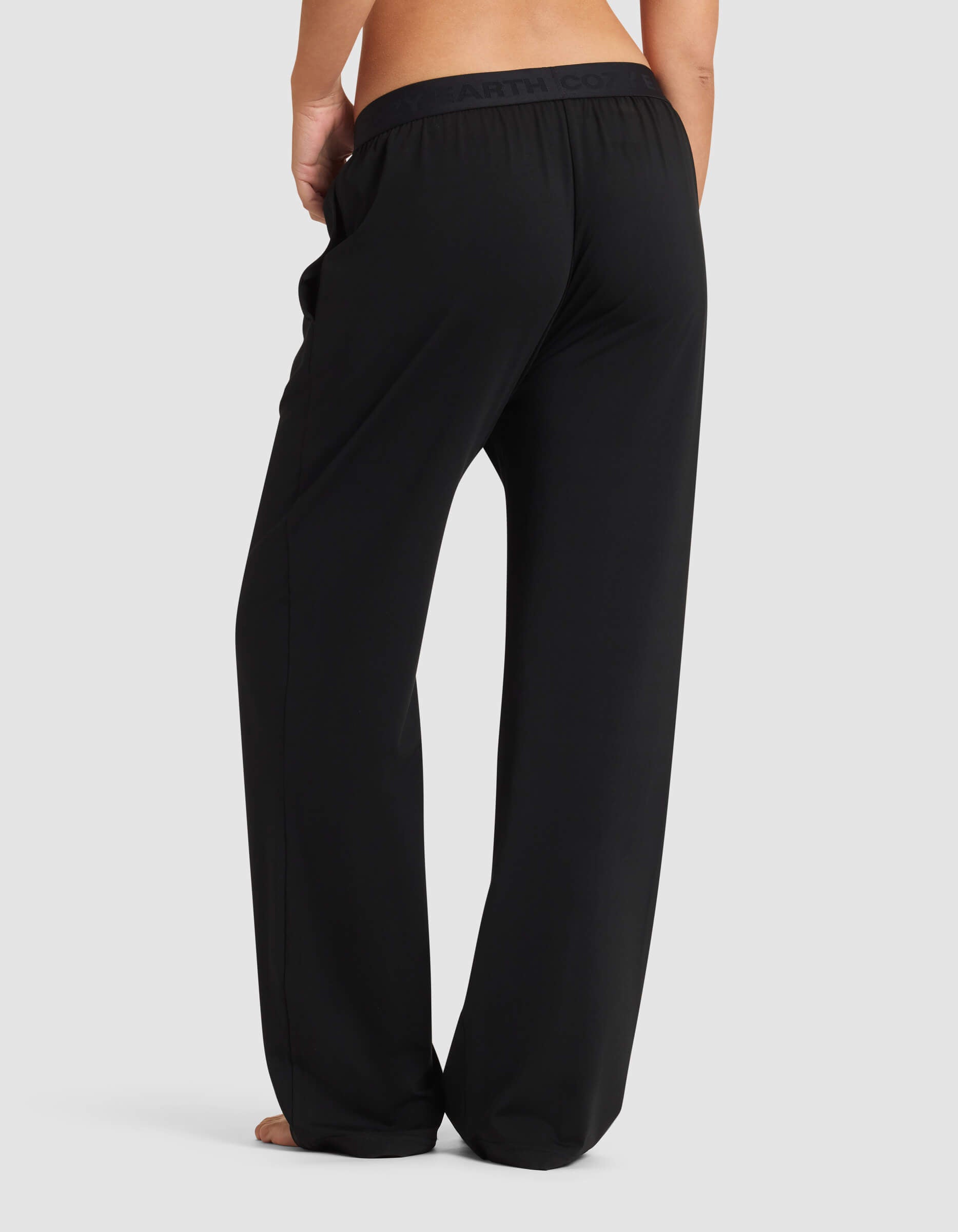 A person stands barefoot, shown from behind, wearing loose-fitting black Cozy Earth Women's First Layer Pajama Pants with hands in the side pockets and a subtle darker waistband.