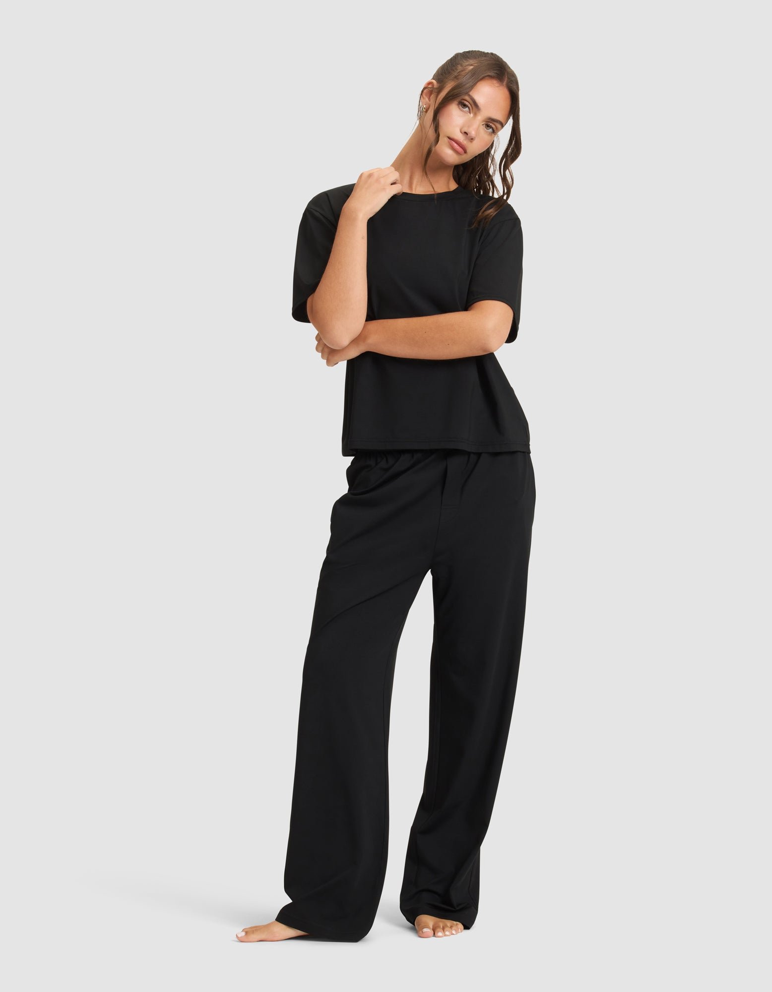 A woman stands barefoot against a plain background, wearing Cozy Earth's Women's First Layer Pajama Pant in black with a loose short-sleeve top, striking a relaxed pose with one hand on her shoulder.
