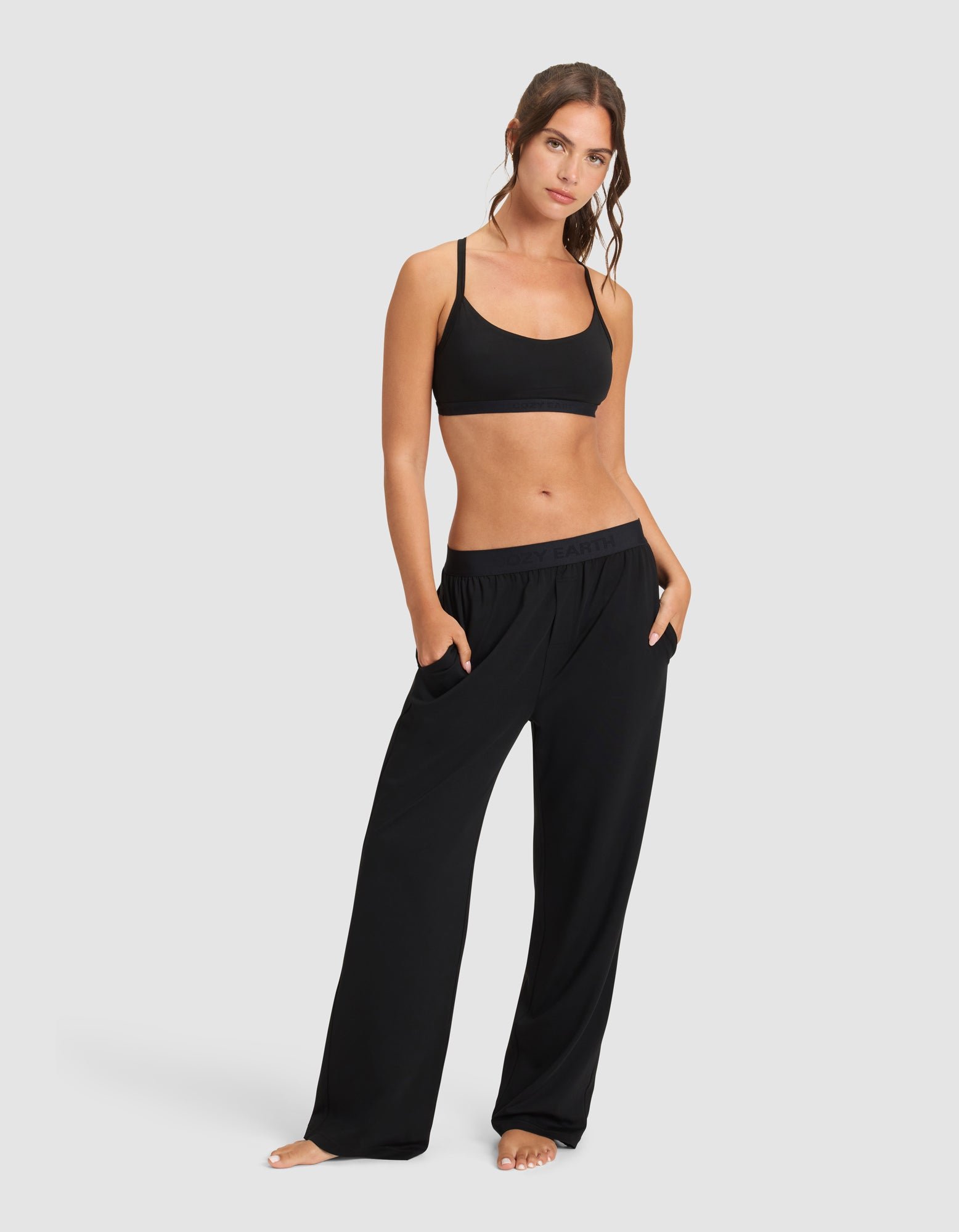 A woman stands barefoot against a plain background, wearing Cozy Earth’s Women's First Layer Pajama Pant in black with her hands in the pockets, paired with a black sports bra, looking at the camera.|Color:Jet Black
