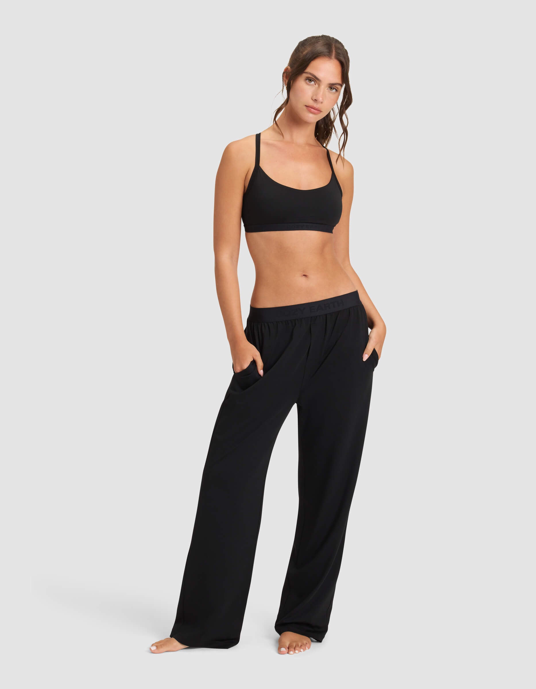A woman stands barefoot against a plain background, wearing Cozy Earth's Women's First Layer Pajama Soft Bra & Pant Set in black, with her hands in the pockets.