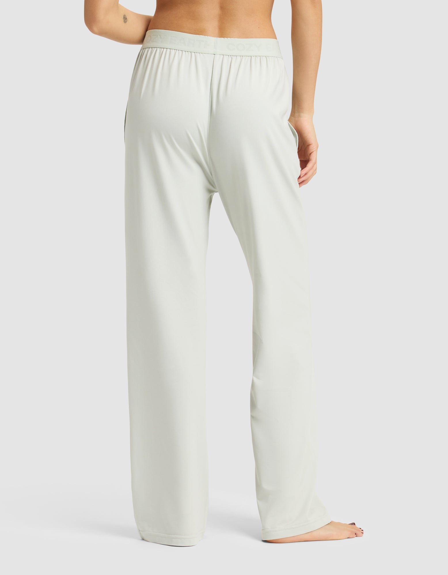 A woman stands barefoot on a white background, seen from the back, wearing Cozy Earth's Women's First Layer Pajama Pant in a light, loose fit with one arm slightly bent and visible.