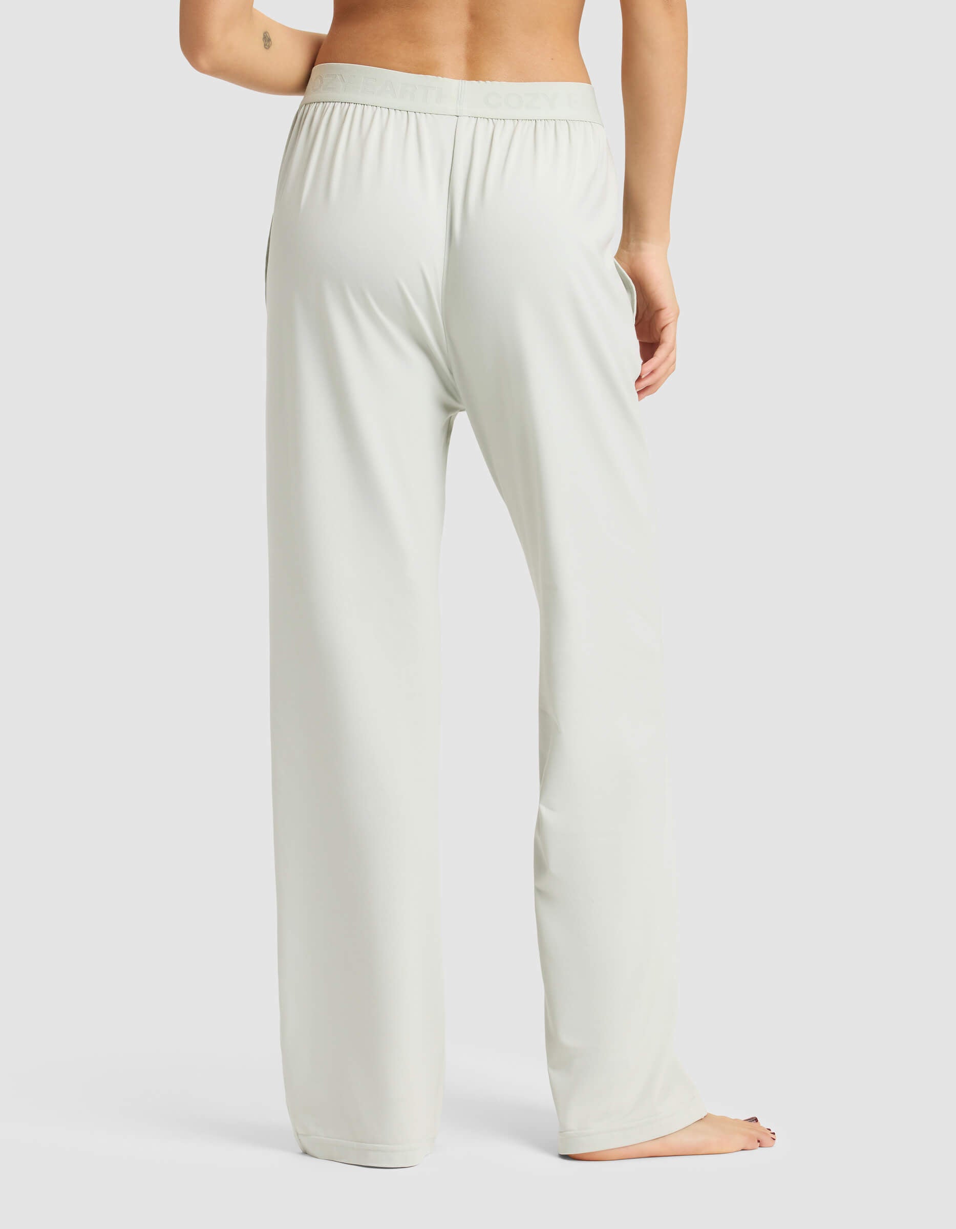 A woman stands barefoot on a white background, seen from the back, wearing Cozy Earth's Women's First Layer Pajama Pant in a light, loose fit with one arm slightly bent and visible.