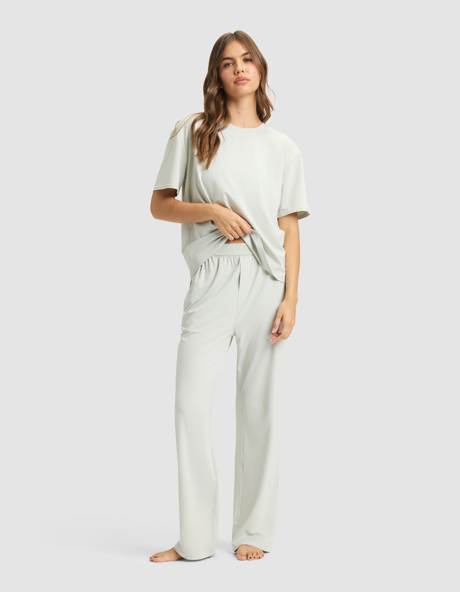 A woman with long, wavy hair stands barefoot against a plain background, wearing Cozy Earth's Women's First Layer Pajama Pant and matching short-sleeve top, both in light colors. She looks relaxed and comfortable.