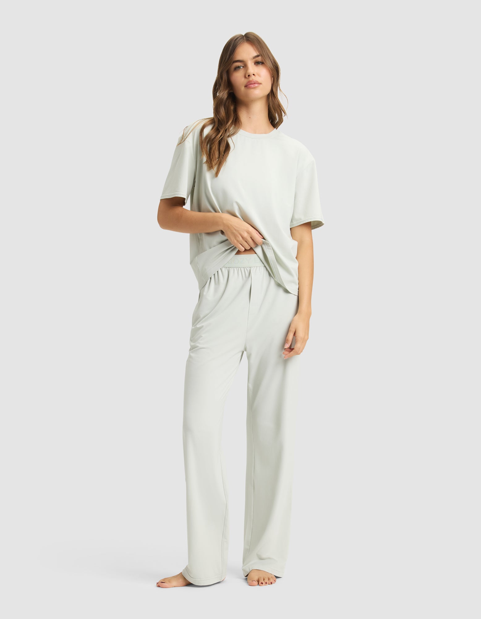A woman with long, wavy hair stands barefoot against a plain background, wearing Cozy Earth's Women's First Layer Pajama Pant and matching short-sleeve top, both in light colors. She looks relaxed and comfortable.