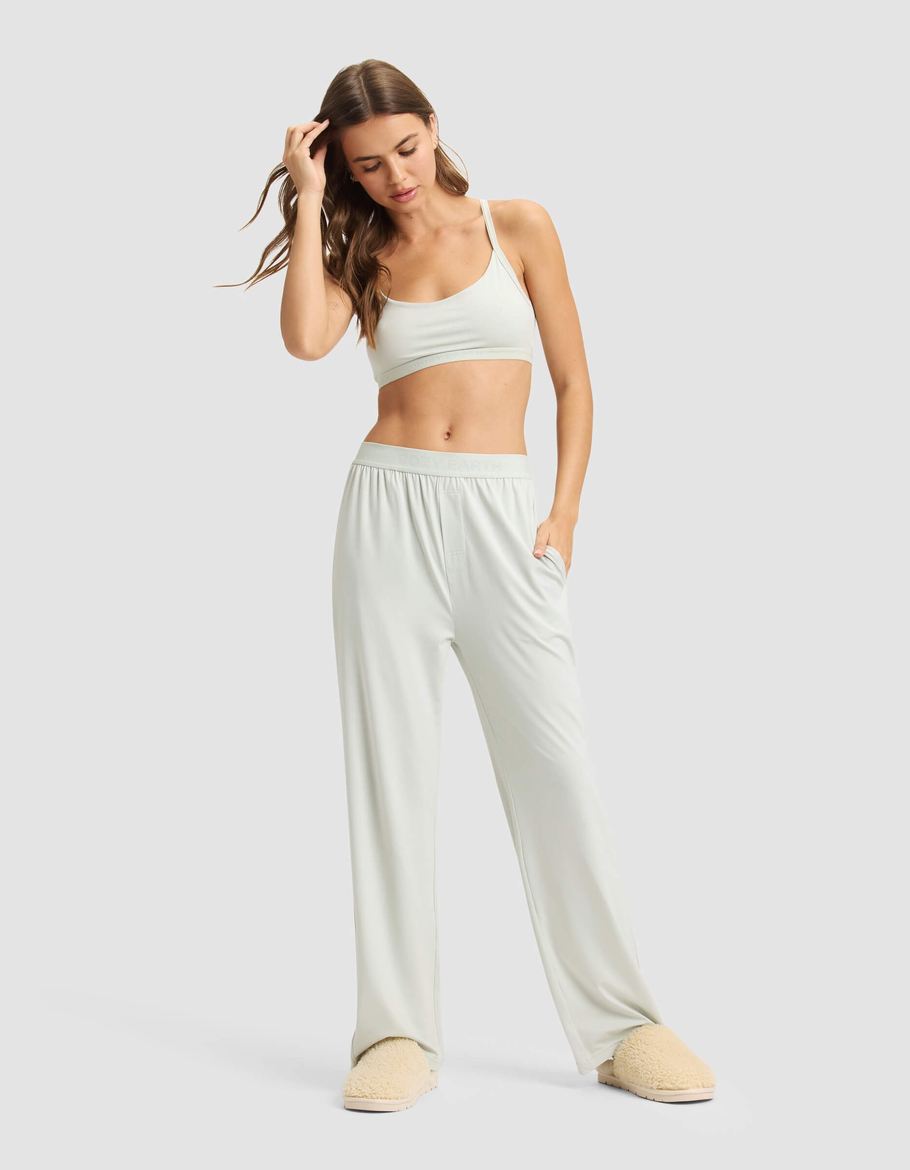 A woman in Cozy Earth's Women's First Layer Pajama Soft Bra & Pant Set stands against a plain background, wearing the light bralette and loose pajama pants with beige slippers, one hand in her pocket and the other touching her hair.