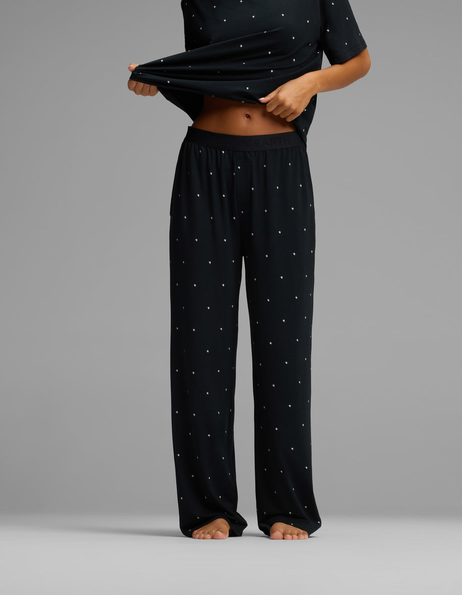 A person stands barefoot against a plain gray background, wearing the Cozy Earth Women’s First Layer Pajama Pant and a matching loose black top with small white polka dots, lifting the shirt slightly to reveal their midriff.