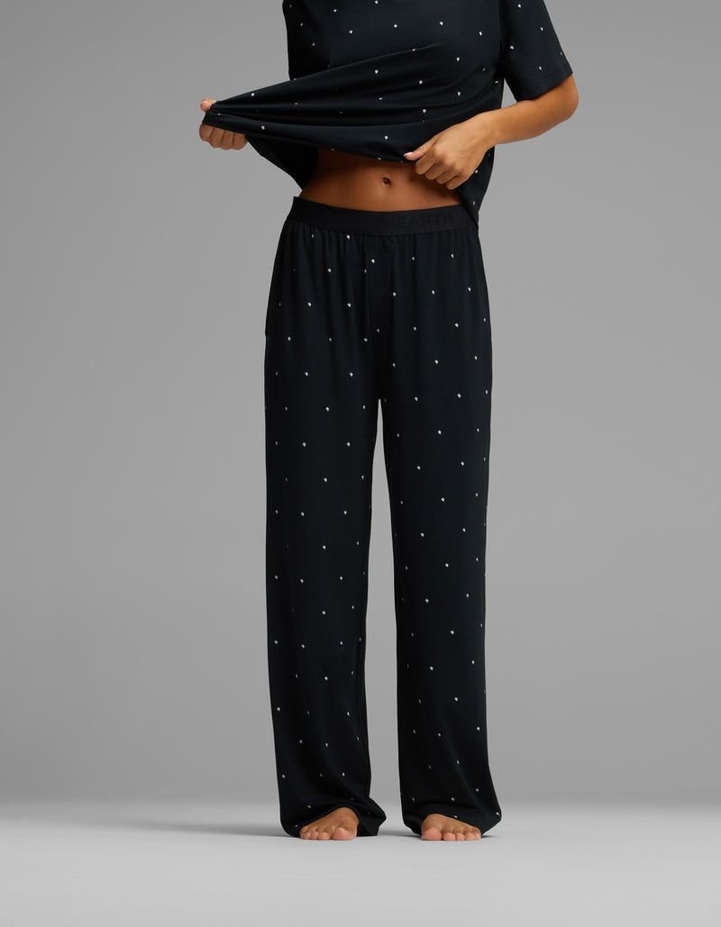 A person stands barefoot against a plain gray background, wearing the Cozy Earth Women’s First Layer Pajama Pant and a matching loose black top with small white polka dots, lifting the shirt slightly to reveal their midriff.