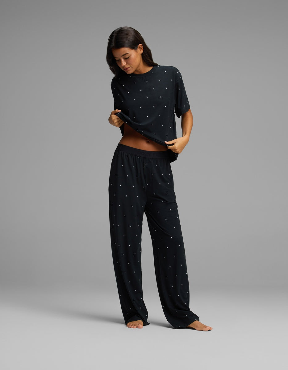 A woman stands barefoot on a gray background, wearing Cozy Earth’s Women’s First Layer Pajama Pant in a black fabric with small white dots. She slightly lifts her shirt to show her stomach while looking down.